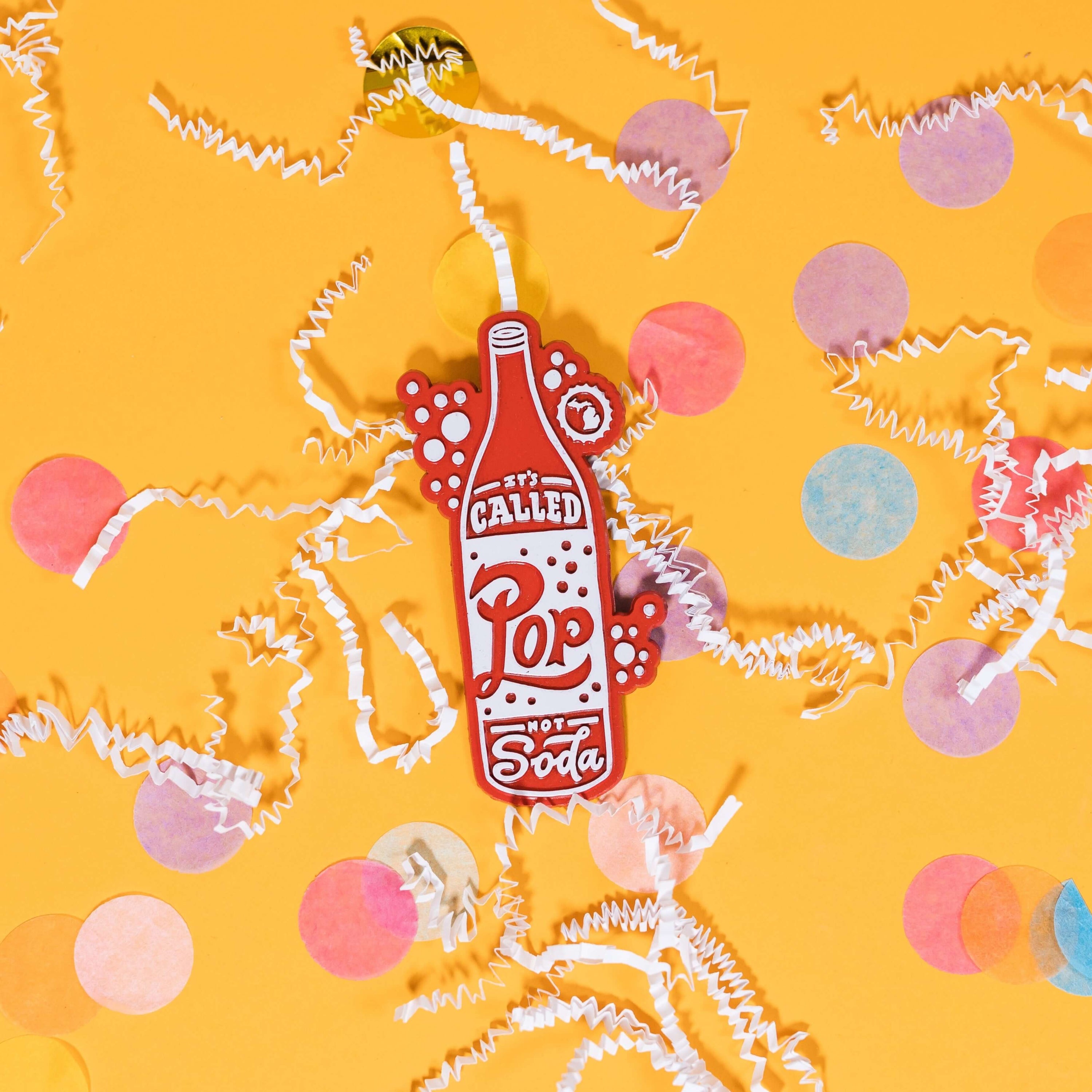 On a sunny mustard background sits a magnet with white crinkle and big, colorful confetti scattered around. This 2D-style raised magnet is a red and white handdrawn soda pop that says "It's Called Pop Not Soda" in red and white handwritten lettering. It has a white outline bubble with the state of Michigan in it in white. 3"- 4"