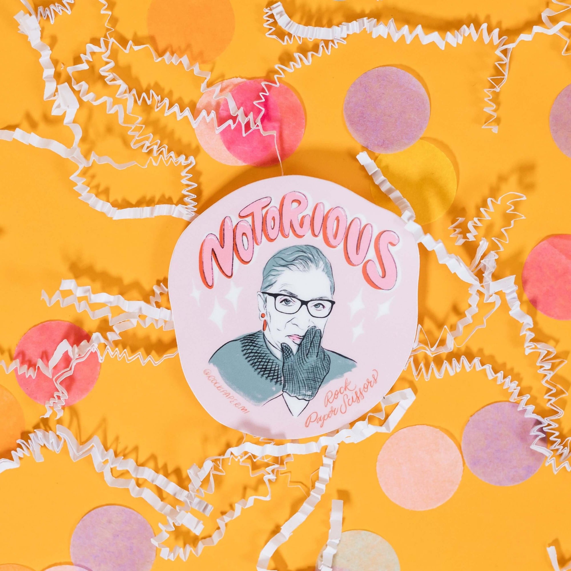 On a sunny mustard background sits a sticker with white crinkle and big, colorful confetti scattered around. This round sticker has an portrait illustration of Ruth Bader Ginsburg on a pink background that says "Notorious" in a pink handdrawn lettering with a red dropshadow. It also says on the bottom "@rockpapermi" in red handwritten lettering and "Rock Paper Scissors" in red handwritten script lettering. 2"x3"