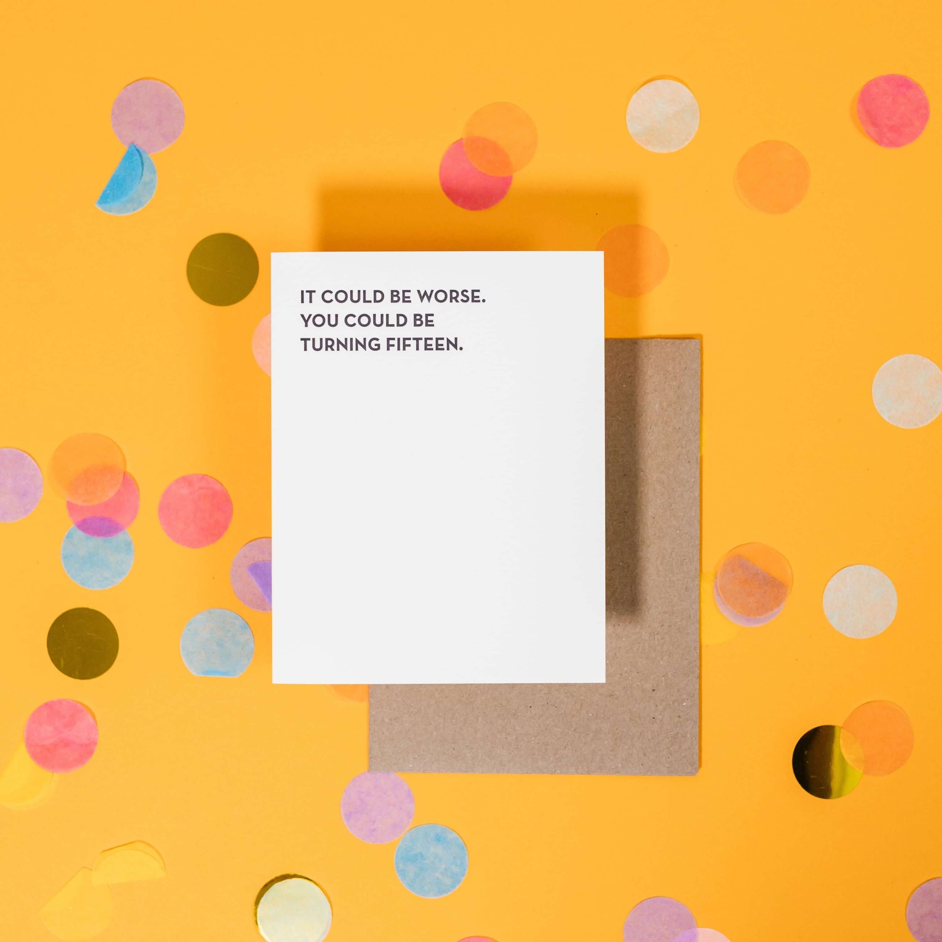 On a sunny mustard background is a greeting card and envelope with big, colorful confetti scattered around. The white greeting card says "It Could Be Worse. You Could Be Turning Fifteen." in a black, sans serif uppercase font. The kraft envelope sits under the card.