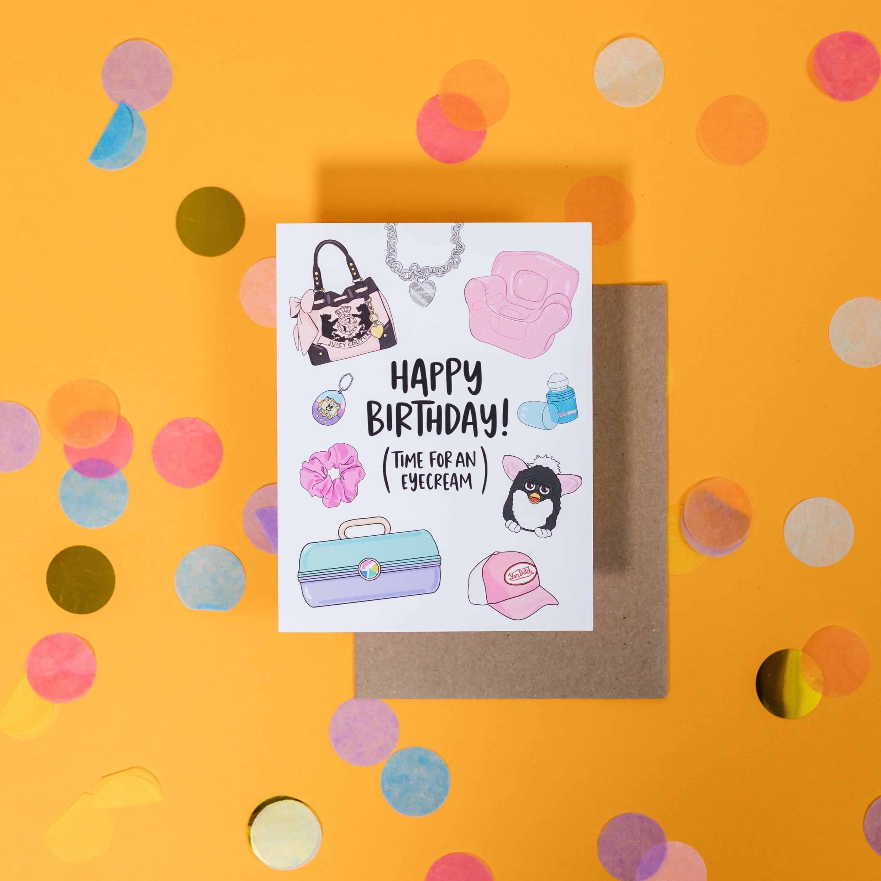 On a sunny mustard background is a greeting card and envelope with big, colorful confetti scattered around. The white greeting card has illustrations from the 90's and says "Happy Birthday (Time For An Eyecream)." The illustrations are Juicy Couture purse, Limited Too heart necklace, pink blow up chair, a body glitter roller, Furby stuffed animal, pink Von Dutch hat, mint and lavender Caboodle, pink Scrunchie, and a Tamagotchi Interactive Virtual Pet. The kraft envelope sits under the card.