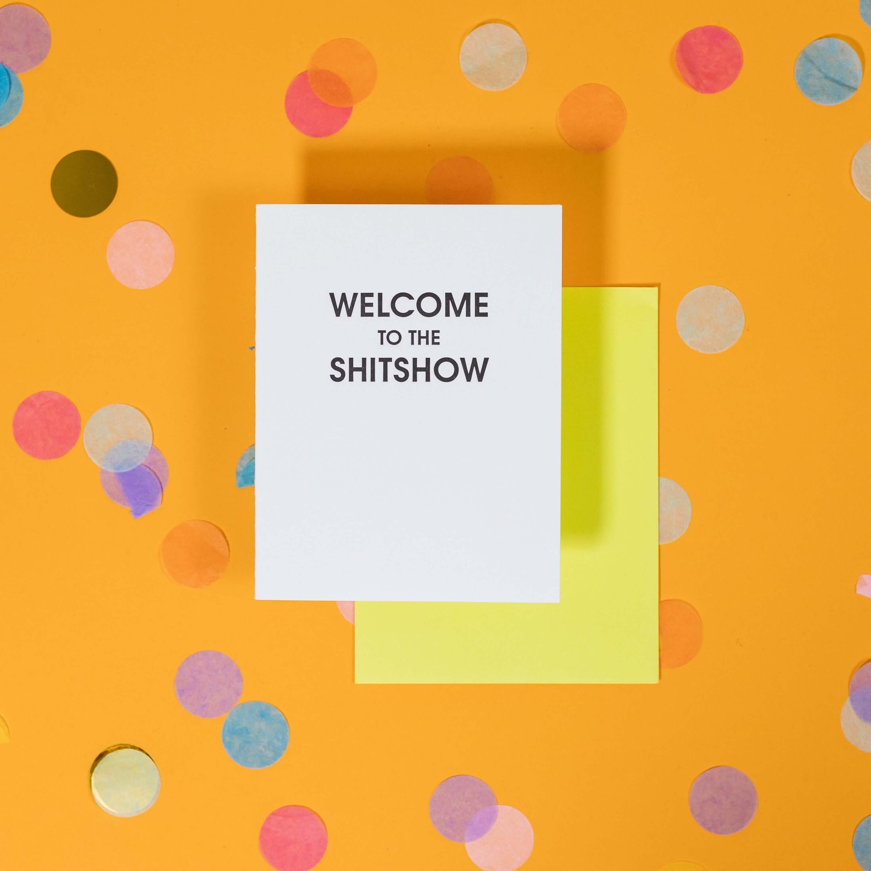 On a sunny mustard background is a greeting card and envelope with big, colorful confetti scattered around. The white greeting card says "WELCOME TO THE SHITSHOW" in big black, sans serif uppercase font. The neon yellow envelope sits under the card.