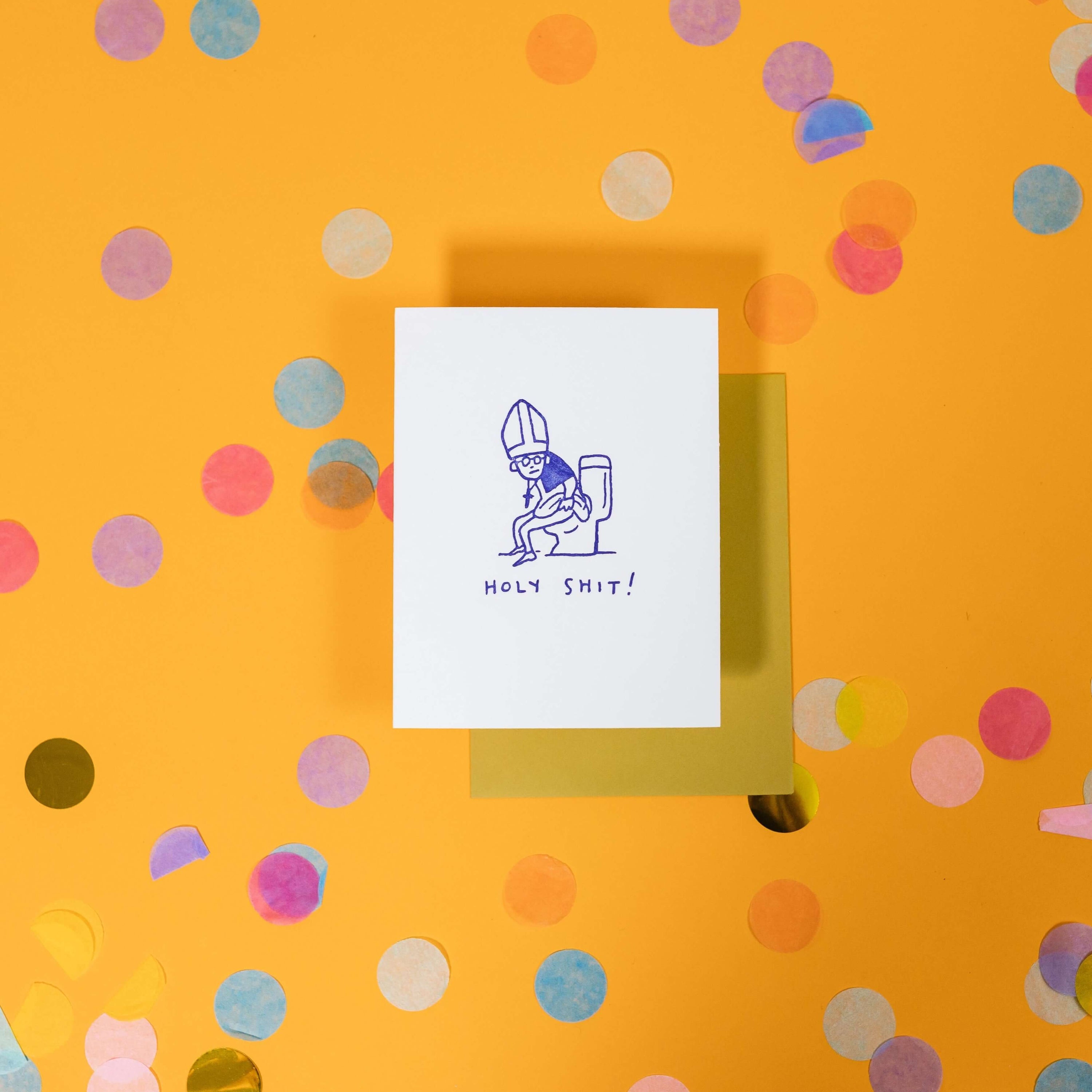On a sunny mustard background is a greeting card and envelope with big, colorful confetti scattered around. The white greeting card has an illustration of a pope sitting on the toilet and it says "HOLY SHIT!" in handwritten purple lettering. The chartreuse envelope sits under the card.