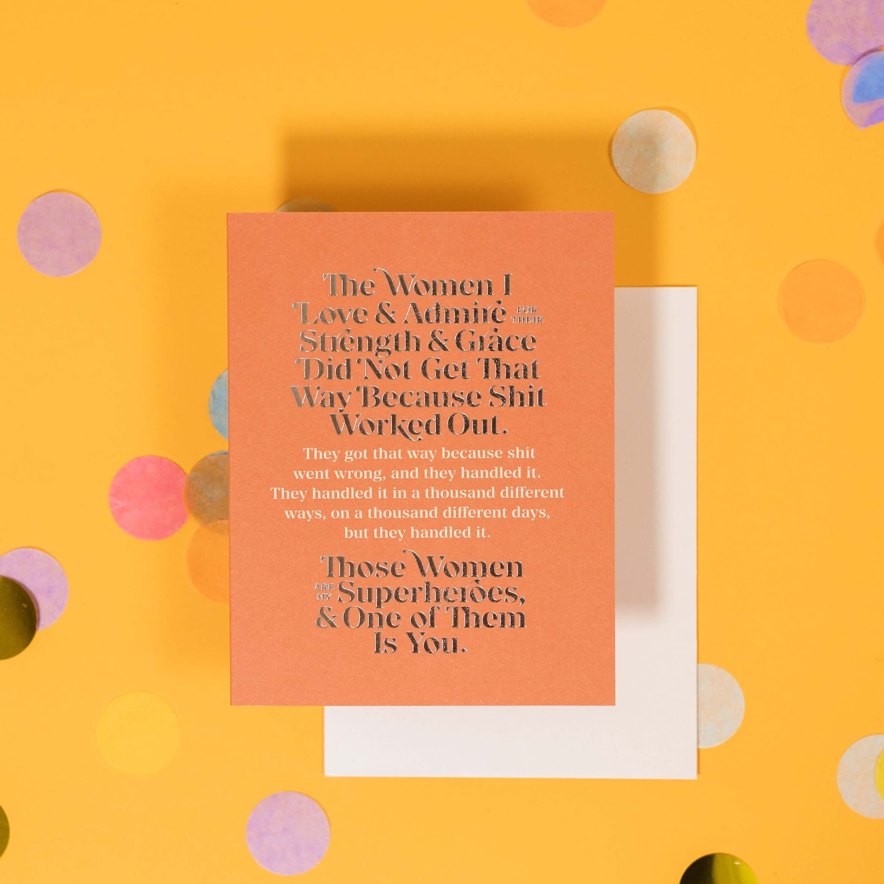 On a sunny mustard background is a greeting card and envelope with big, colorful confetti scattered around. The orange greeting card says "The Women I Love & Admire For Their Strength & Grace Did Not Get That Way Because Shit Worked Out. Those Women Are My Superheroes, & One of Them Is You." It is written in gold foil and white serif font. The white envelope sits under the card.