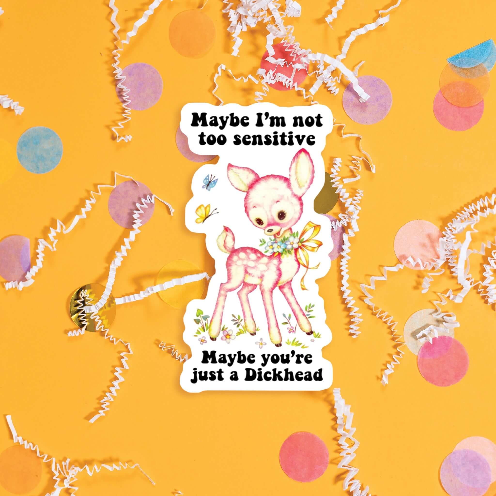 On a sunny mustard background sits a sticker with colorful confetti and white crinkle scattered around. This vintage sticker has an illustration of a cute pink deer with a floral garland tied with a yellow ribbon around the neck. It says "Maybe I'm not too sensitive Maybe you're just a Dickhead" in black, thick serif font. Approximately 3".