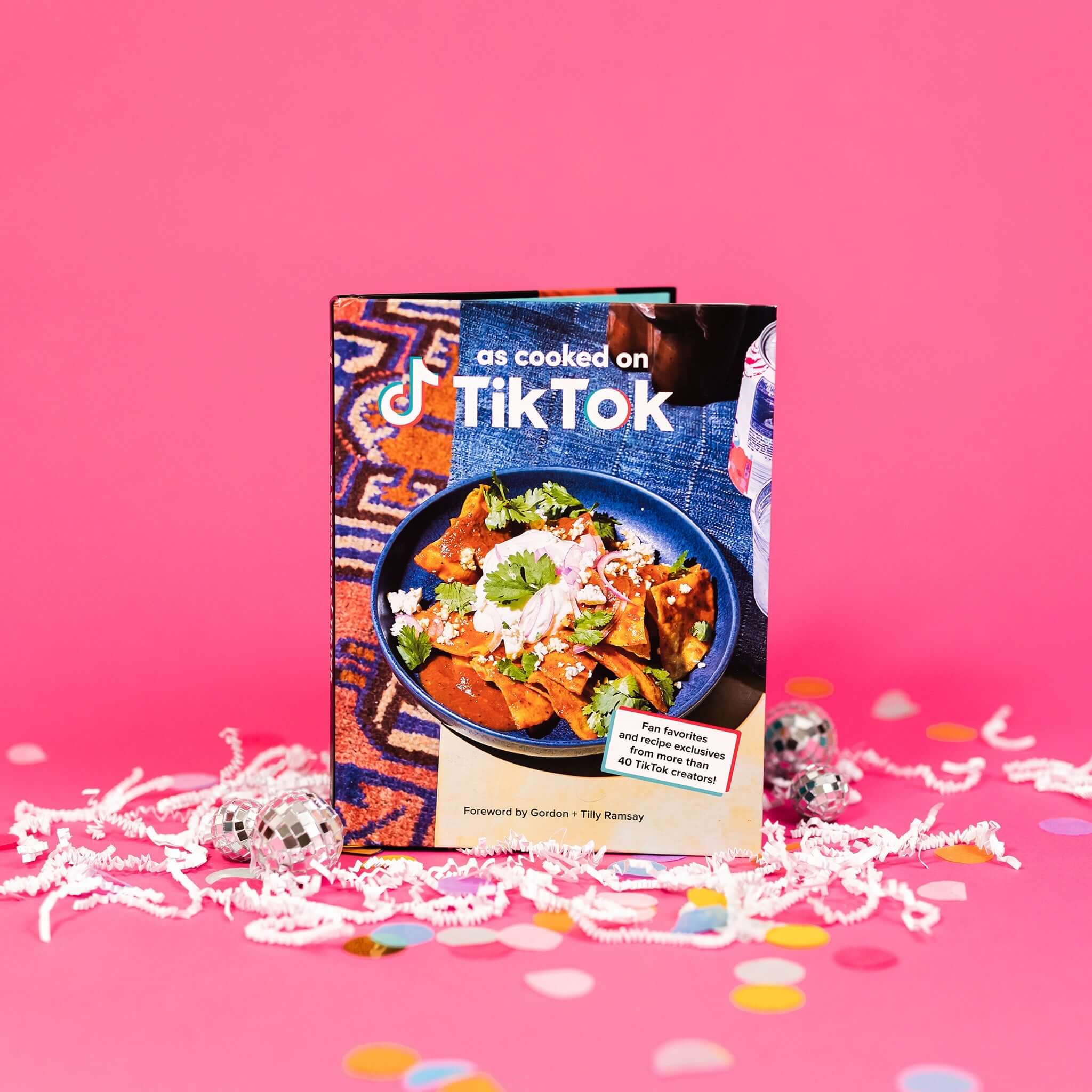 On hot pink background sits a book with white crinkle and big, colorful confetti. There are mini disco balls. This book has a picture of a blue bowl filled with tortilla chips, cilantro, white cheese and onions. It says "as cooked on TikTok" in a white, block font. In a white box it says "Fan favorites and recipe exclusives from more than 40 TikTok creators!" with a red and turquoise border. It also says "Foreword by Gordon + Tilly Ramsay" in black, block font.