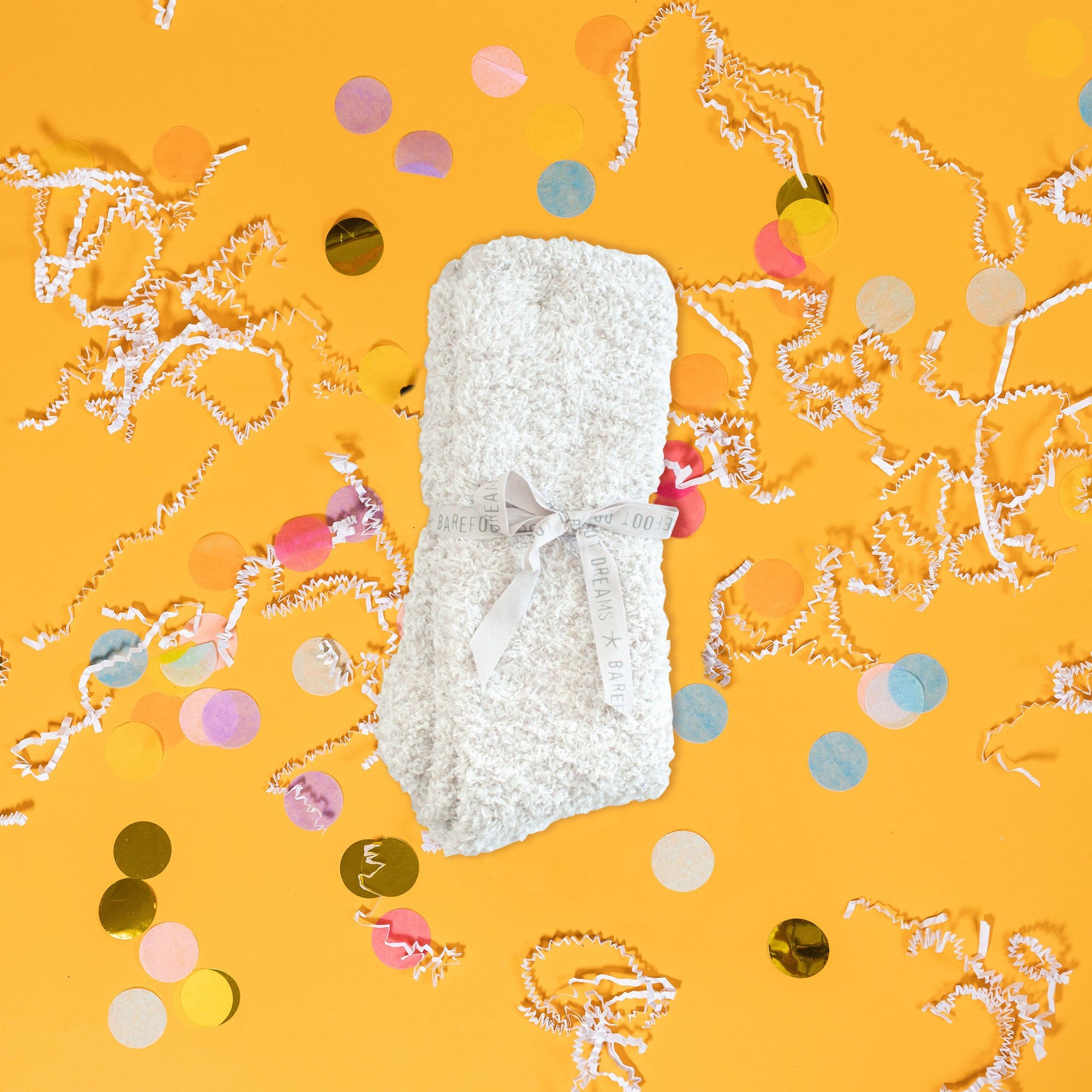 On a sunny mustard background sits a pair of socks folded and tied with a light stone ribbon that says "BAREFOOT DREAMS" repeatedly. These heathered socks are stone and white and so soft. There are white crinkle and big, colorful confetti scattered around.