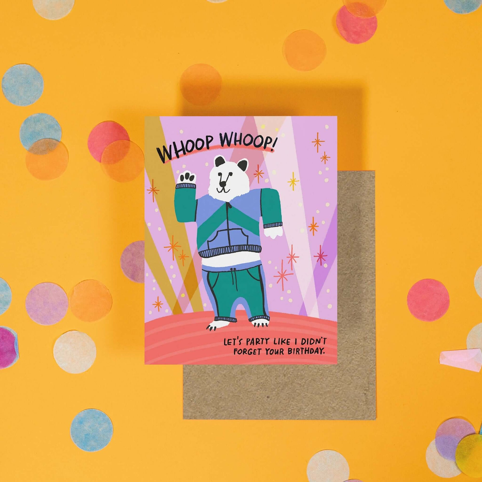 On a sunny mustard background is a greeting card and envelope with big, colorful confetti scattered around. The greeting card has an colorful illustration of a bear in a warm up suit and it says at the top "WHOOP WHOOP!" in a black, handwritten lettering. It also says "Let's party like I didn't forget your birthday." in a black, handwritten lettering. The kraft envelope sits under the card.