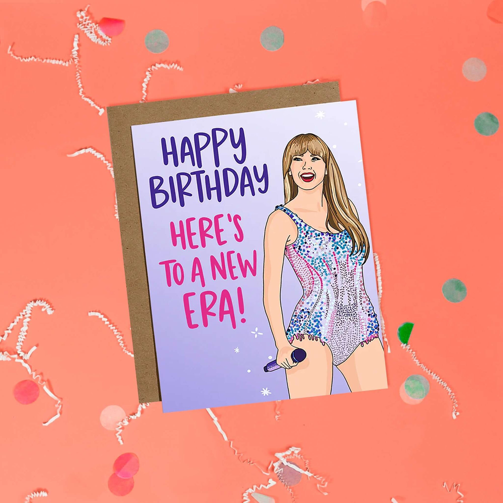 On a coral pink background sits a card with white crinkle and big, colorful confetti scattered around. This Taylor Swift inspired card has different shades of lavender. There is an illustration of Taylor Swift and it says "HAPPY BIRTHDAY HERE'S TO A NEW ERA!" in purple and pink lettering. It has a kraft envelope.