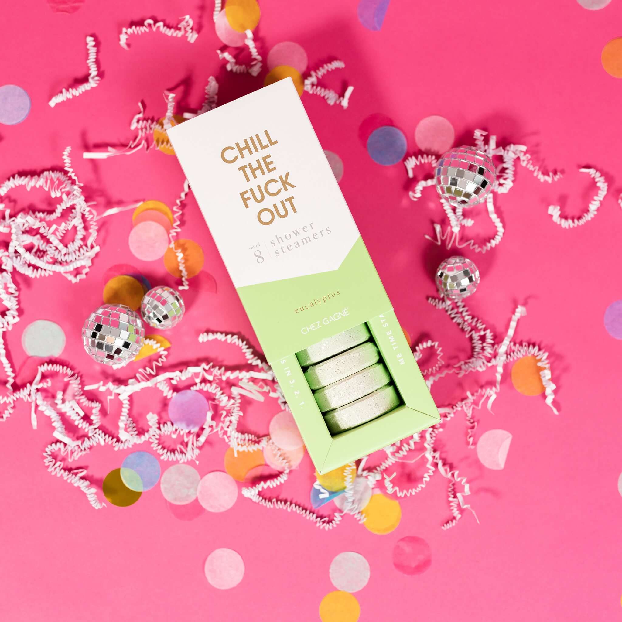 On a hot pink background sits an opened box with white crinkle and big, colorful confetti scattered around. There are mini disco balls. This picture is a close-up of a white and pistachio green package that says "CHILL THE FUCK OUT" in gold foil, all caps block lettering. Under it says "set of 8" and " shower steamers" in grey, lowercase serif font. At the bottom it says "eucalyptus" in gold foil, lower case serif font. The box is opened to reveal the pistachio green shower steamers in it.
