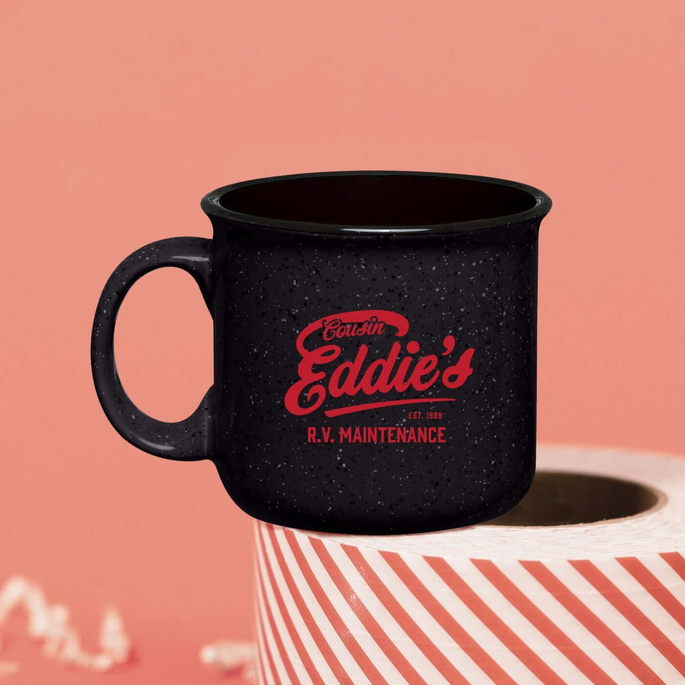 On a pink coral background sits a campfire mug on a roll of red and white packing tape. There is white crinkle. This Christmas Vacation inspired mug is charcoal grey with black and white specks and it's say in red lettering "Cousin Eddie's R.V. MAINTENANCE EST. 1989."