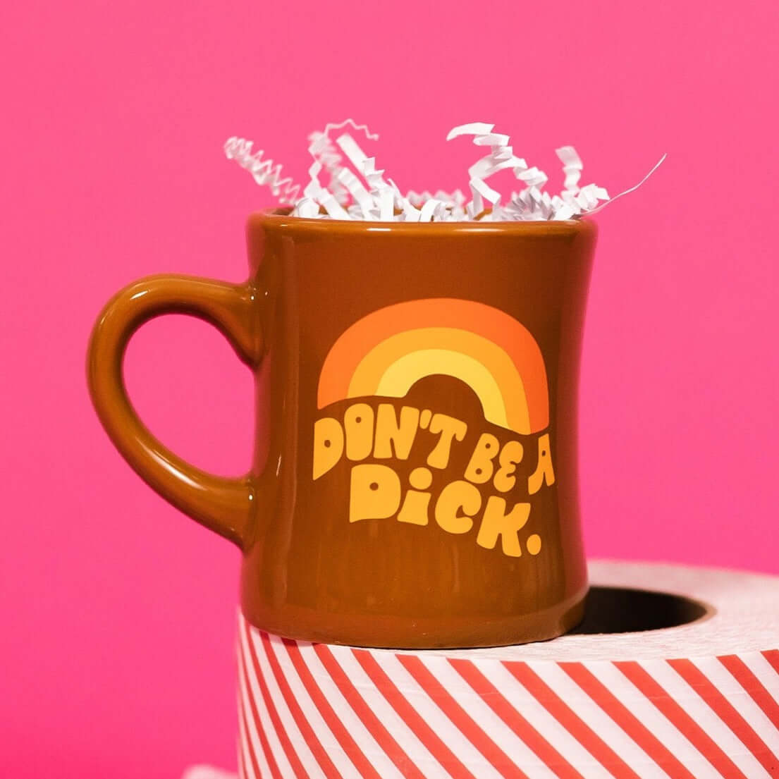 On a hot pink background sits a mug with white crinkle and big, colorful confetti scattered around. The mug is a brown diner style mug with orange, groovy style font that says "Don't Be A Dick." with an orange, light orange and yellow rainbow above it. It has white crinkle in it and sits atop a red and white striped roll of tape.