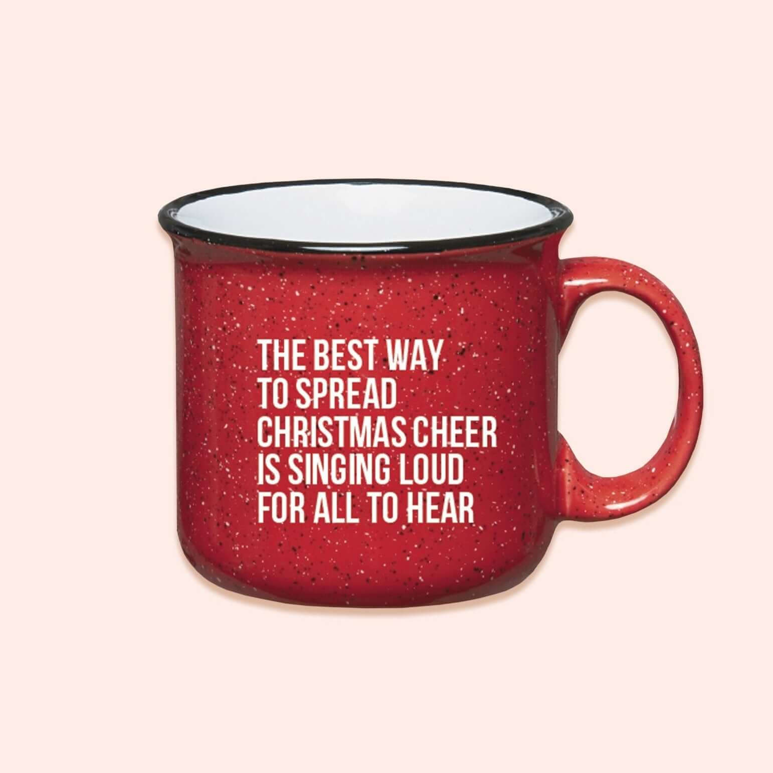 On a light pink background sits a campfire mug. This Elf inspired mug is red with black and white specks and it's say in white lettering "THE BEST WAY TO SPREAD CHRISTMAS CHEER IS SINGING LOUD FOR ALL TO HEAR."