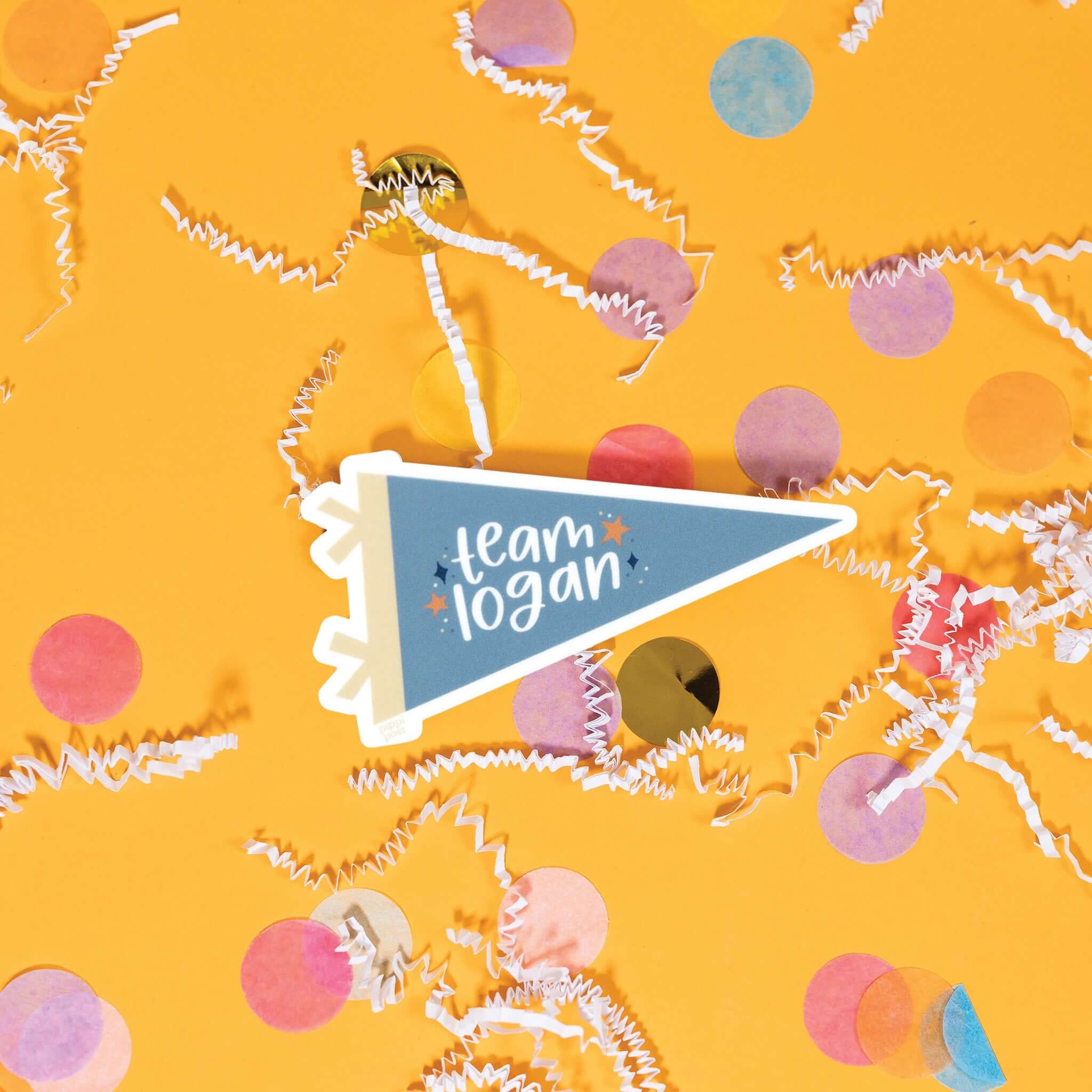 On a sunny mustard background is a sticker with white crinkle and big, colorful confetti scattered around. This Gilmore girls inspired vintage banner shaped sticker is new blue and khaki and has navy and orange stars. It says "team logan" in white, lower case handwritten lettering. 3"x1.6"