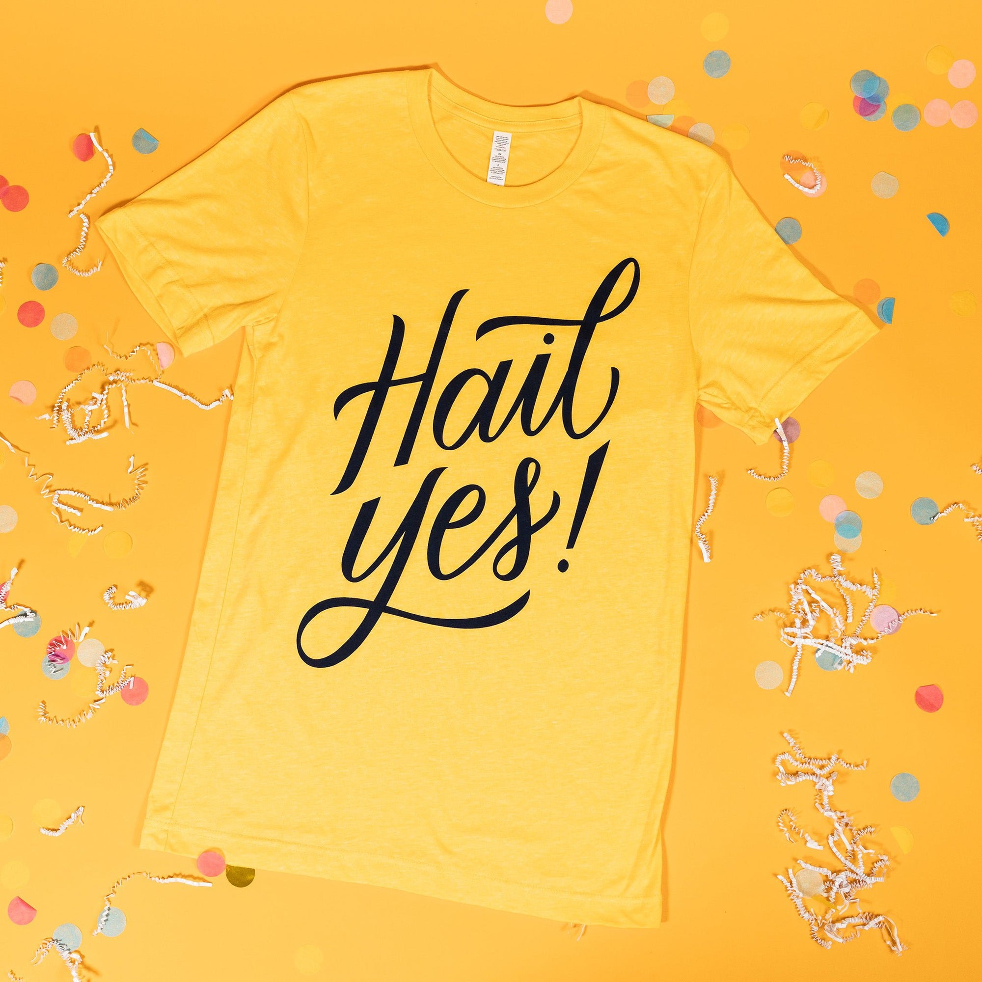 On a sunny mustard background sits a t-shirt with white crinkle and big, colorful confetti scattered around. This yellow tee says "Hail Yes!" in blue lettering.