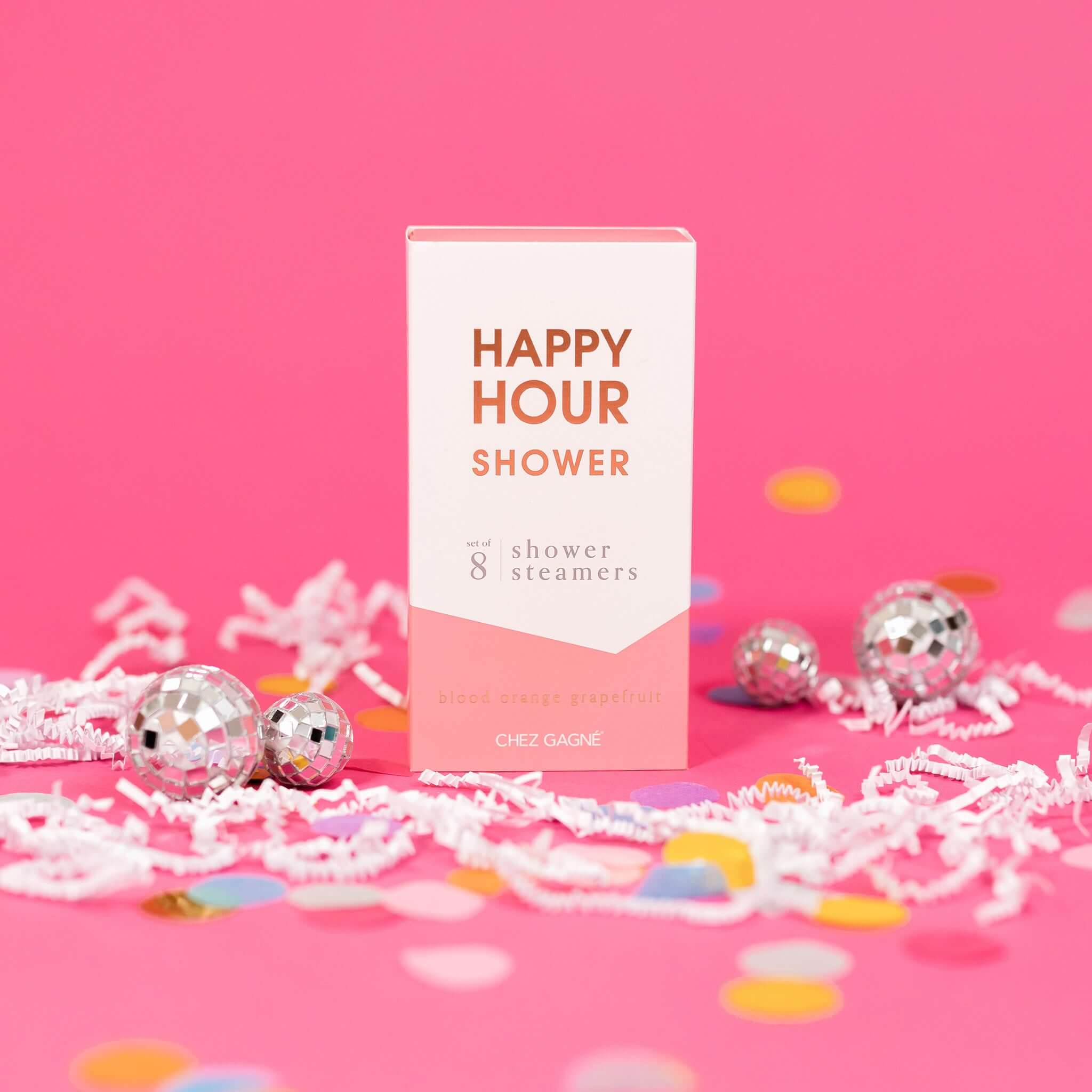 On a hot pink background sits a box with white crinkle and big, colorful confetti scattered around. There are mini disco balls. This picture is a close-up of a white and coral package that says "HAPPY HOUR SHOWER" in gold foil, all caps block lettering. Under it says "set of 8" and " shower steamers" in grey, lowercase serif font. At the bottom it says "blood orange grapefruit" in gold foil, lower case serif font.