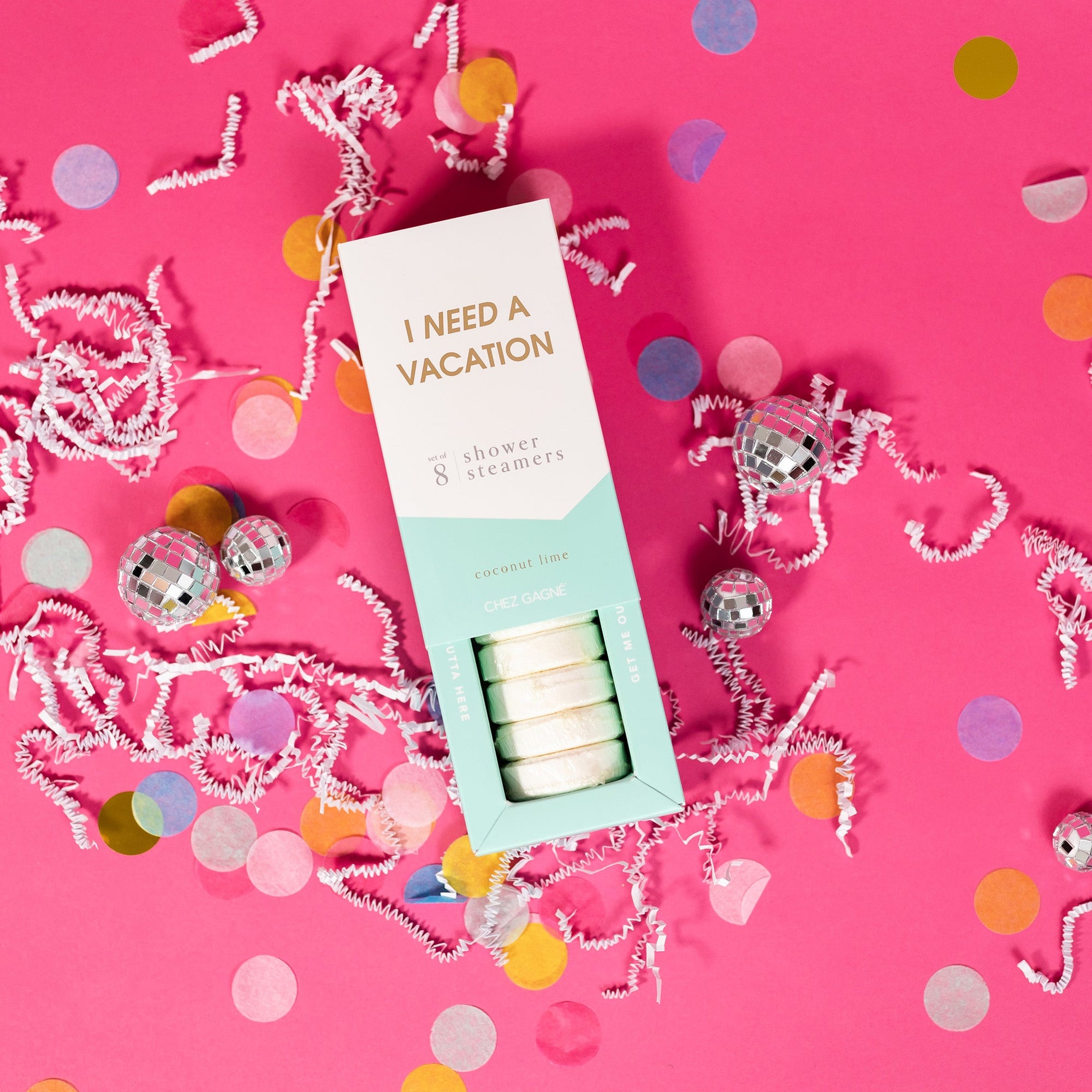 On a hot pink background sits an opened box with white crinkle and big, colorful confetti scattered around. There are mini disco balls. This picture is a close-up of a white and pool blue package that says "I NEED A VACATION" in gold foil, all caps block lettering. Under it says "set of 8" and " shower steamers" in grey, lowercase serif font. At the bottom it says "coconut lime" in gold foil, lower case serif font. The box is opened to reveal the white shower steamers in it. 