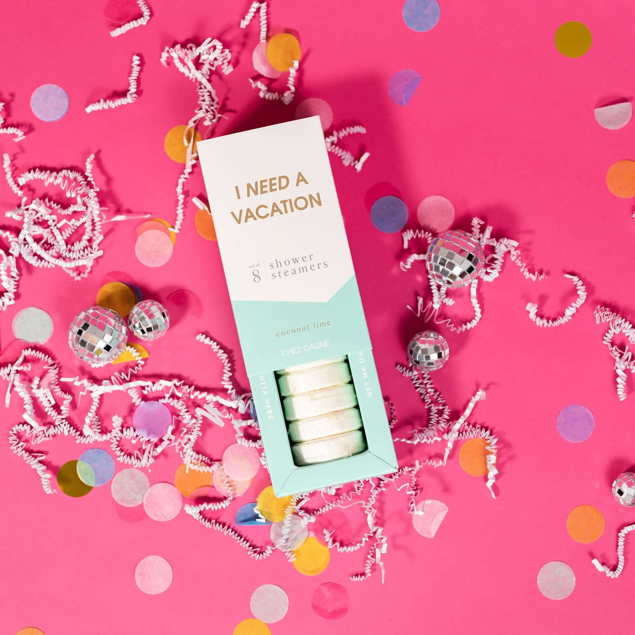 On a hot pink background sits an opened box with white crinkle and big, colorful confetti scattered around. There are mini disco balls. This picture is a close-up of a white and pool blue package that says "I NEED A VACATION" in gold foil, all caps block lettering. Under it says "set of 8" and " shower steamers" in grey, lowercase serif font. At the bottom it says "coconut lime" in gold foil, lower case serif font. The box is opened to reveal the white shower steamers in it.