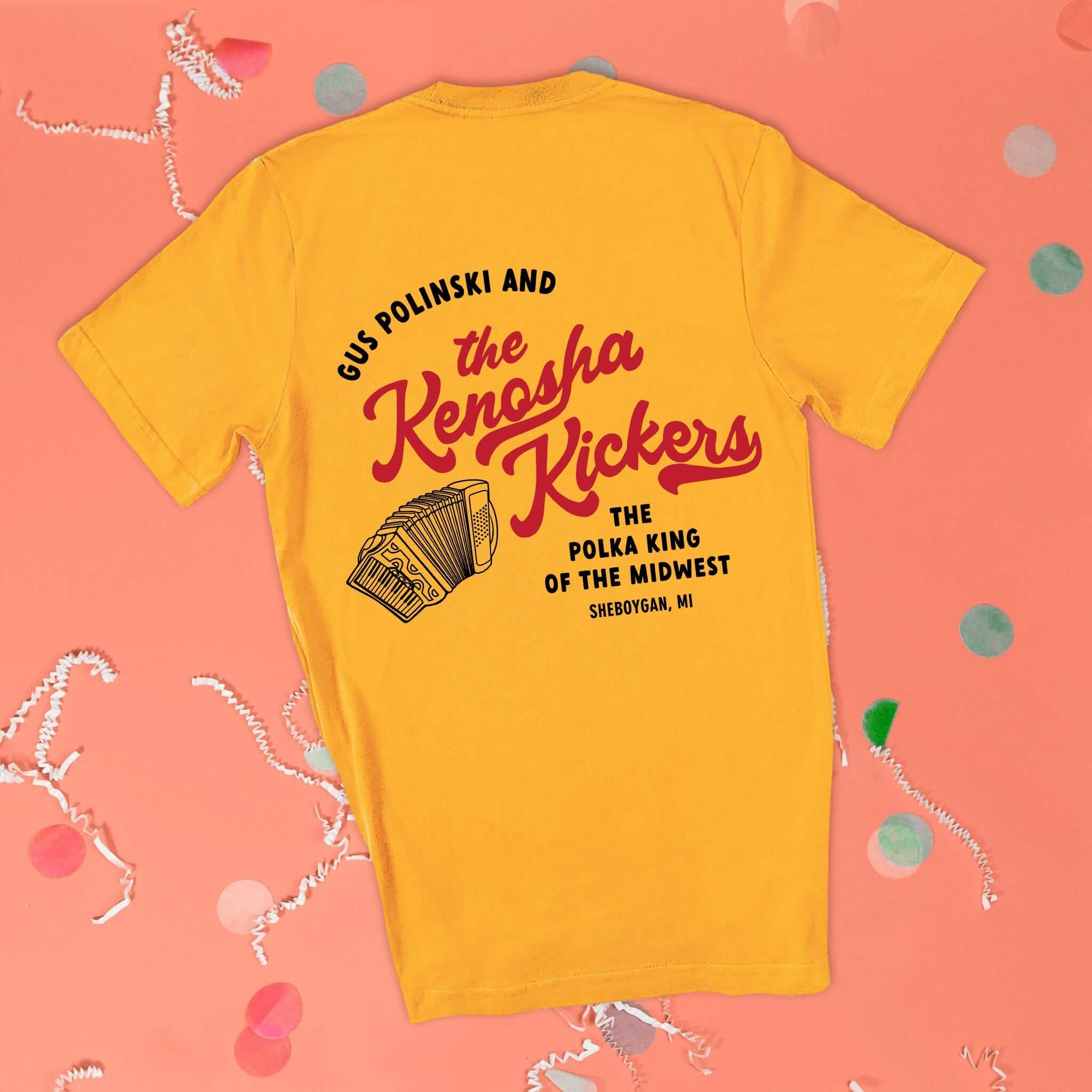On a pink coral background sits the back of a t-shirt with white crinkle and big, colorful confetti scattered around. This Home Alone Inspired crewneck sunny mustard t-shirt has red script lettering black lettering. There is an illustration of an accordian and it says "GUS POLINSKI AND the Kenosha Kickers THE POLKA KIN OF THE MIDWEST SHEBOYGAN, MI."