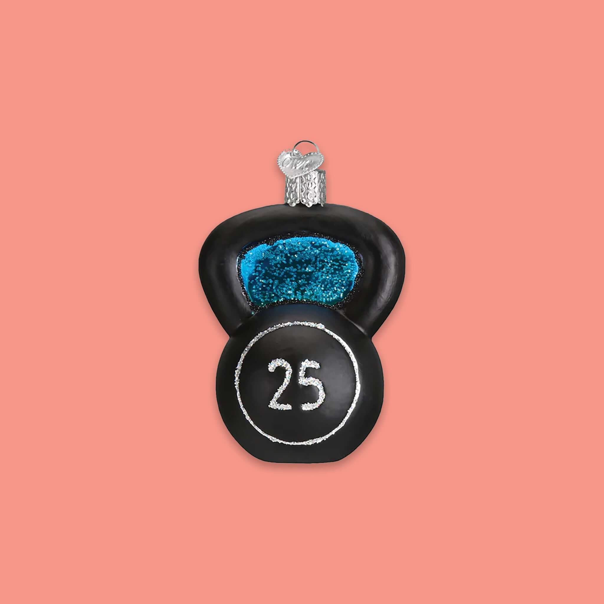 On a coral pink background sits an ornament. This glass ornament is of a black kettlebell with a silver glitter circle and the number '25' in silver glitter inside the circle.