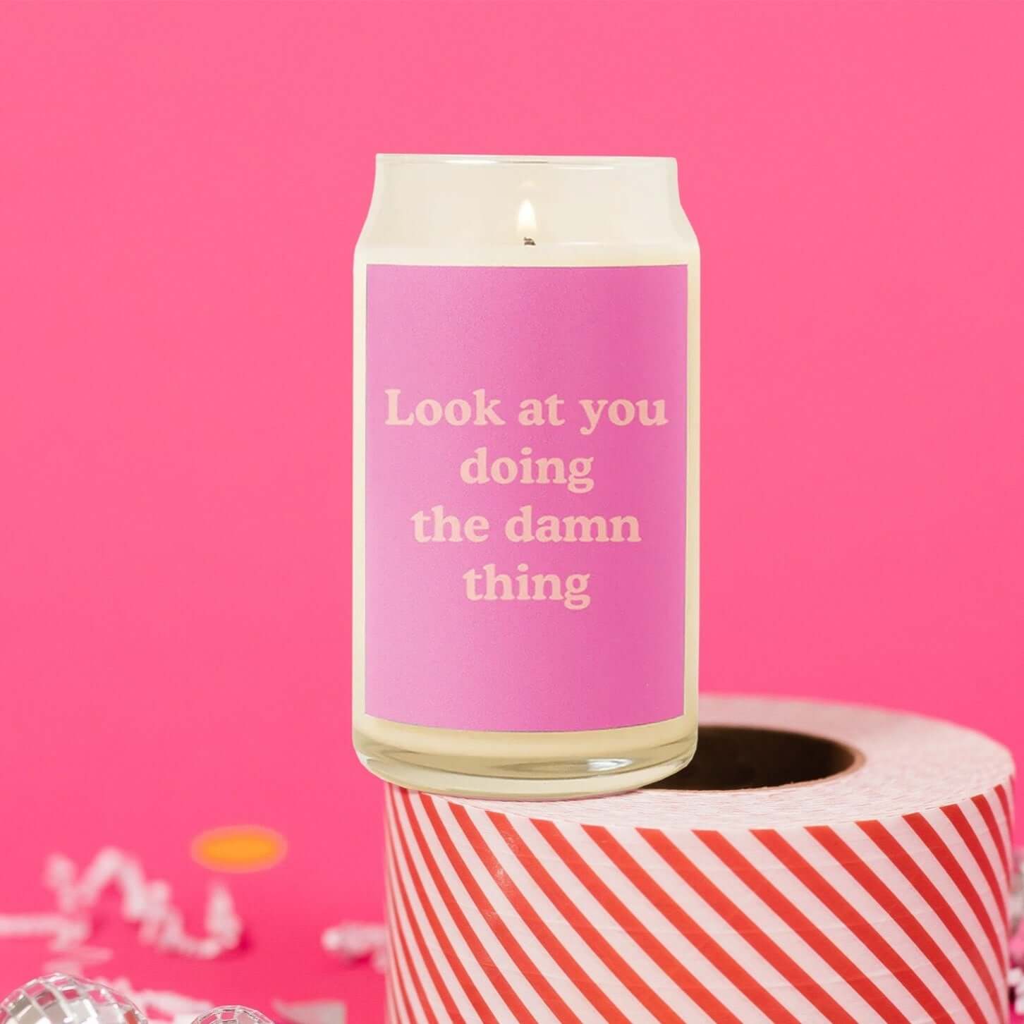 On a hot pink background sits a candle on a roll of red and white striped packing tape with white crinkle and big, colorful confetti scattered around. The glass candle is lit and has a hot pink label on the front and it says "Look at you doing the damn thing" in a coral, thick serif font.