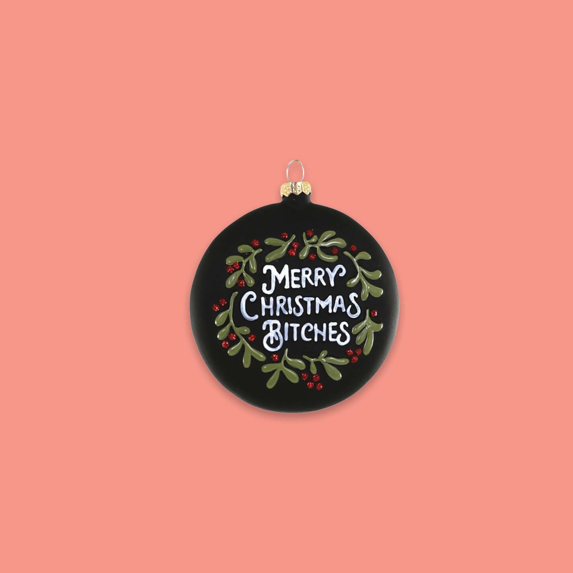 On a coral pink background sits a round ornament. This glass ornament is black with green and red hollies and berries and it says "MERRY CHRISTMAS BITCHES" in white lettering.