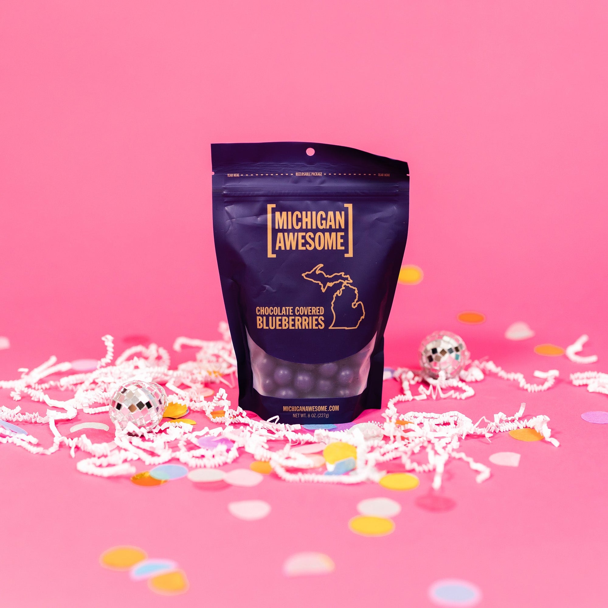 On a bubblegum pink background sits a bag with white crinkle and big, colorful confetti scattered around. There are mini disco balls. It is filled with chocolate covered blueberries. It says "MICHIGAN AWESOME," and "CHOCOLATE COVERED BLUEBERRIES" in goldish-yellow, all caps block font. It has an outlined illustration of the state of Michigan-UP. NET WT. 8 OZ. (227G)