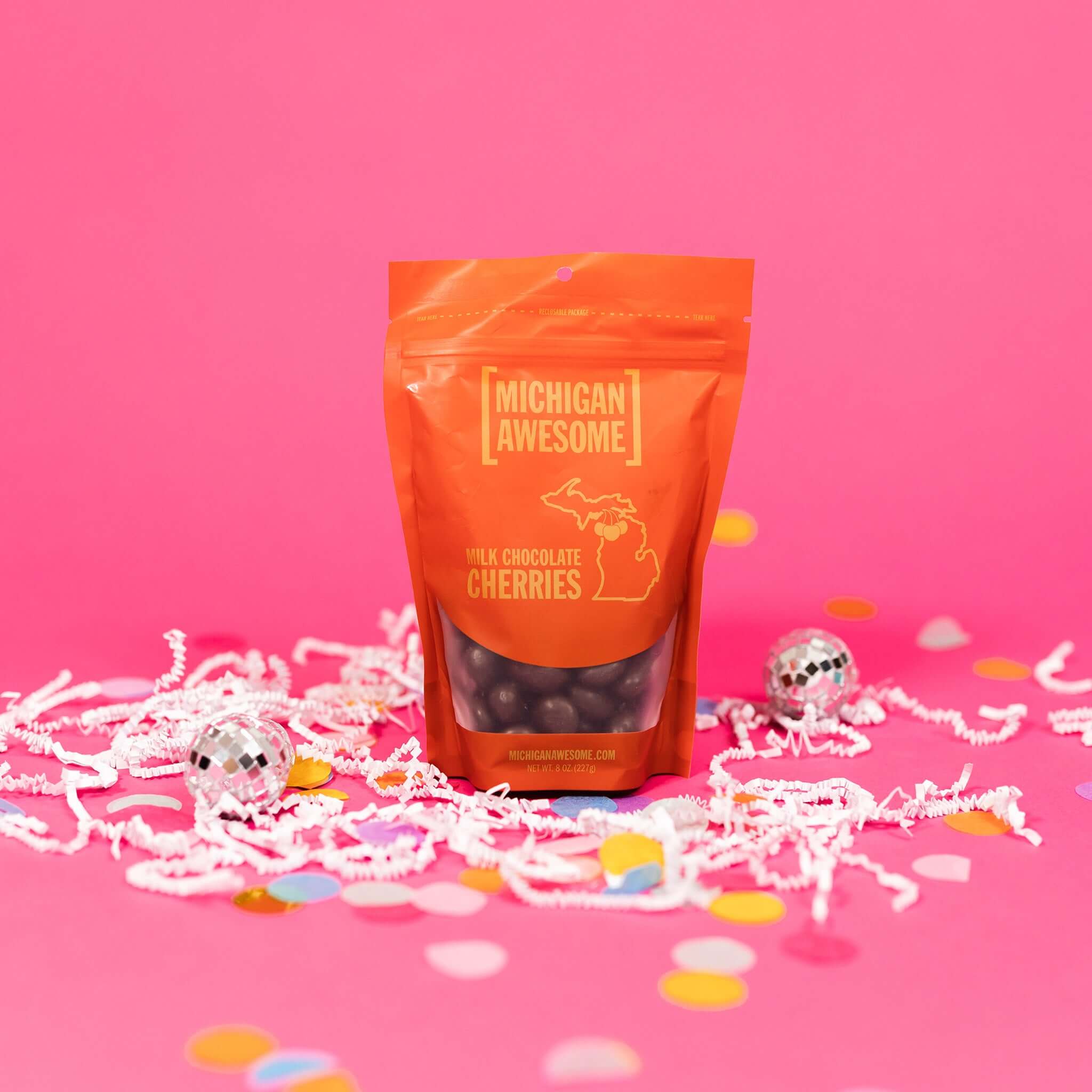 On a bubblegum pink background sits a bag with white crinkle and big, colorful confetti scattered around. There are mini disco balls. This red package has chocolate covered cherries in the package. It says in gold "MICHIGAN AWESOME," "MILK CHOCOLATE CHERRIES" in all caps block font. It also says "MICHIGANAWESOME.COM" in gold, all caps block font. NEW WT. 8 OZ. (227g)