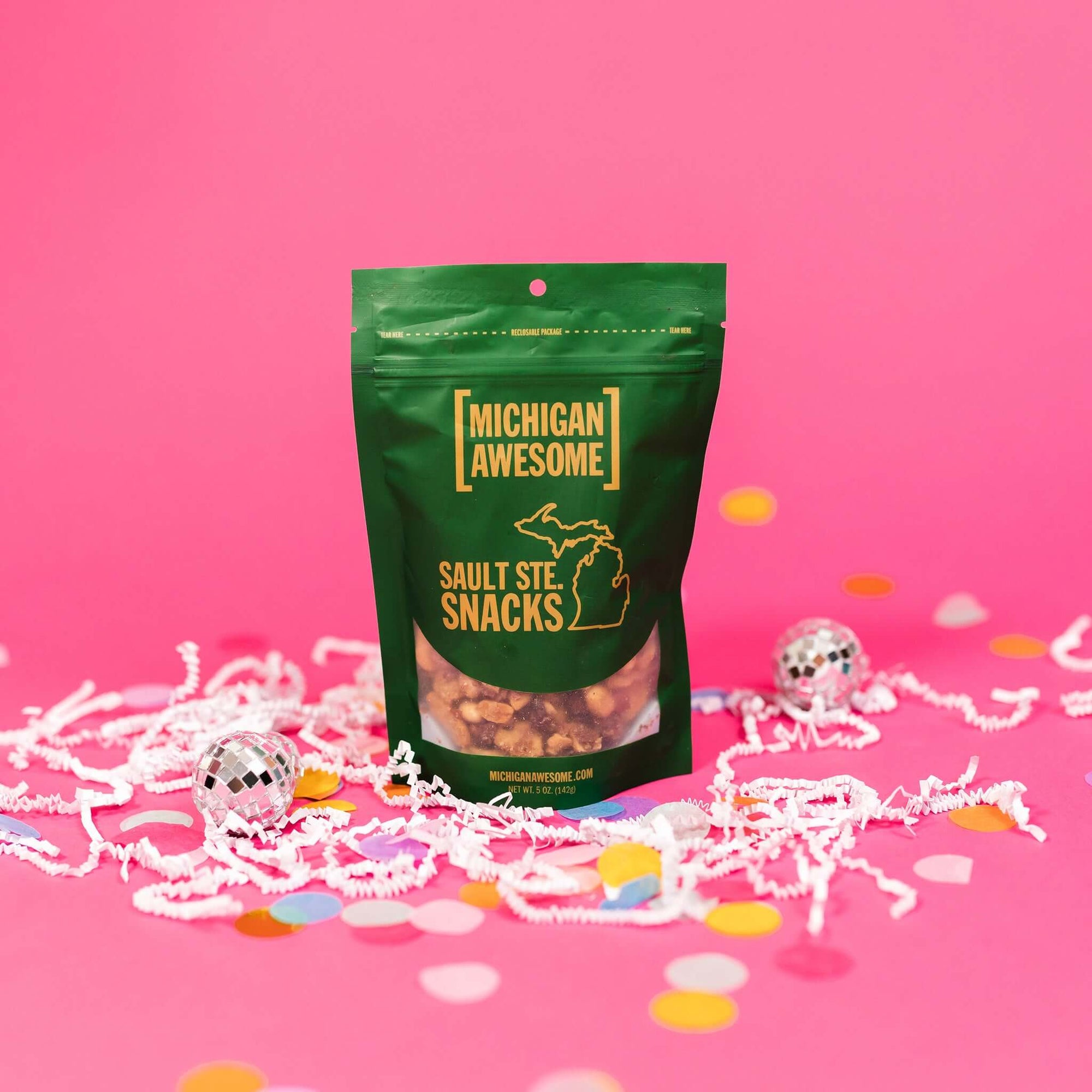 On a bubblegum pink background sits a bag with white crinkle and big, colorful confetti scattered around. There are mini disco balls. This green bag filled with sesame sticks, sesame seeds, and salt and pepper peanuts. It says "MICHIGAN AWESOME," and "SAULT STE. SNACKS" in goldish-yellow, all caps block font. It has an outlined illustration of the state of Michigan-UP.