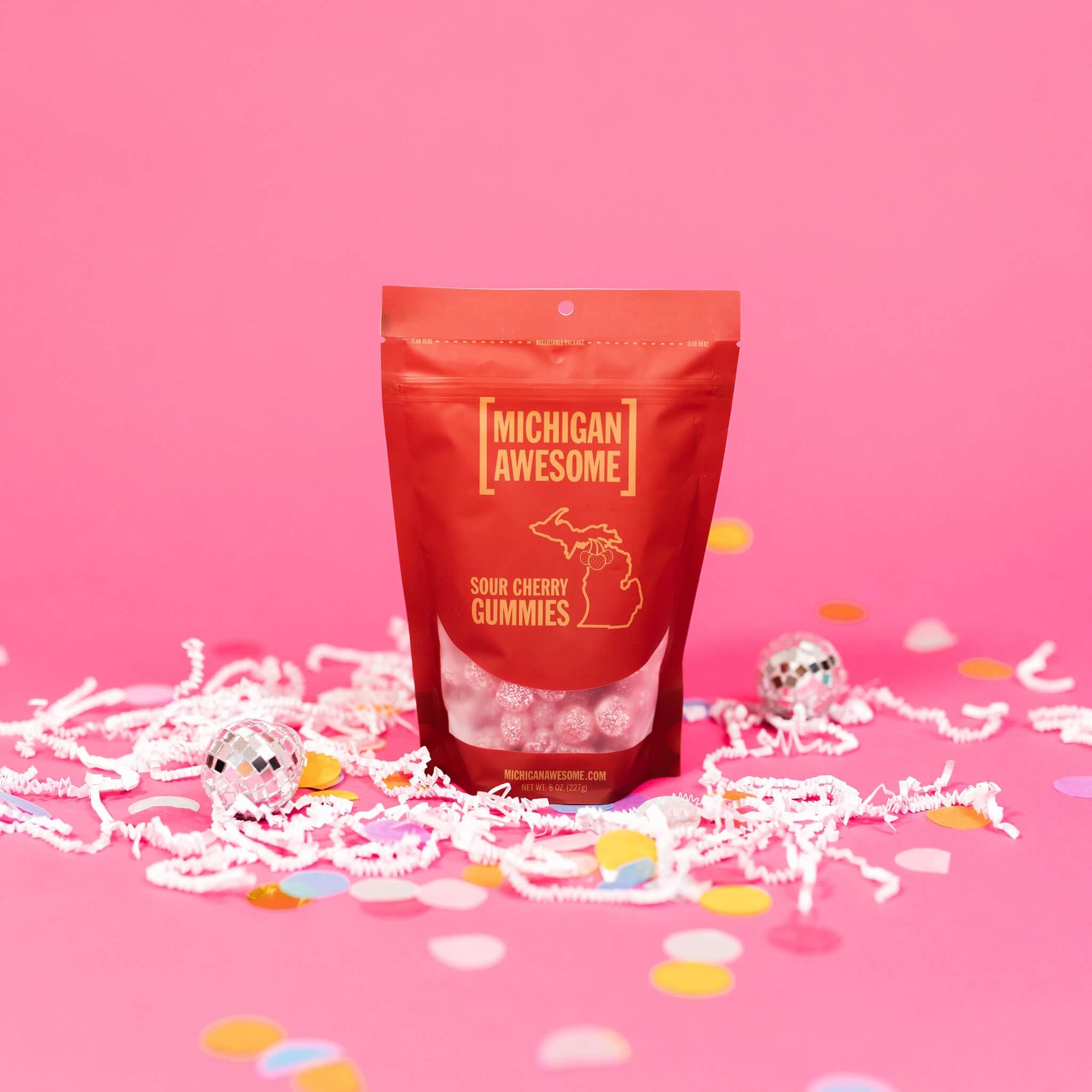 On a bubblegum pink background sits a bag with white crinkle and big, colorful confetti scattered around. There are mini disco balls. This red bag filled with sugar sour cherry gummies. It says "MICHIGAN AWESOME," and "SOUR CHERRY GUMMIES" in goldish-yellow, all caps block font. It has an outlined illustration of the state of Michigan-UP with cherries. There are sugar sour cherry gummies sitting around the bag.