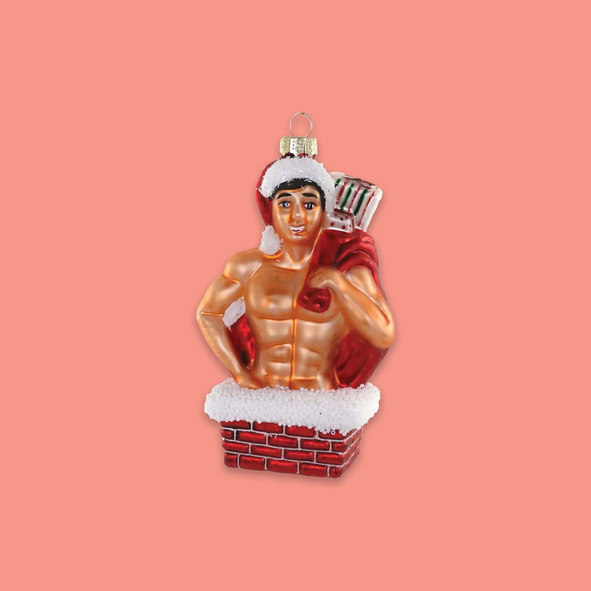 On a coral pink background sits an ornament. This glass ornament is of a naked man with a six pack, wearing a santa hat and holding a santa gift bag. He is inside of a chimney.