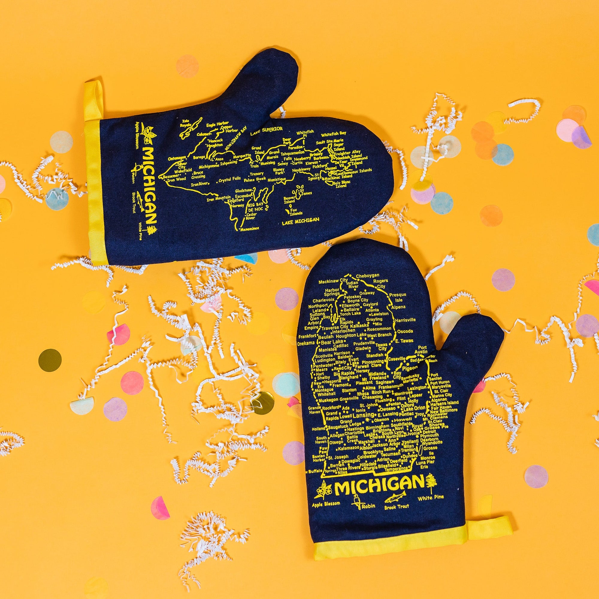 On a sunny mustard background sits a pair of oven mitts with white crinkle and big, colorful confetti scattered around. These navy and yellow oven mitts feature a yellow map of Michigan and the UP with cities in yellow.