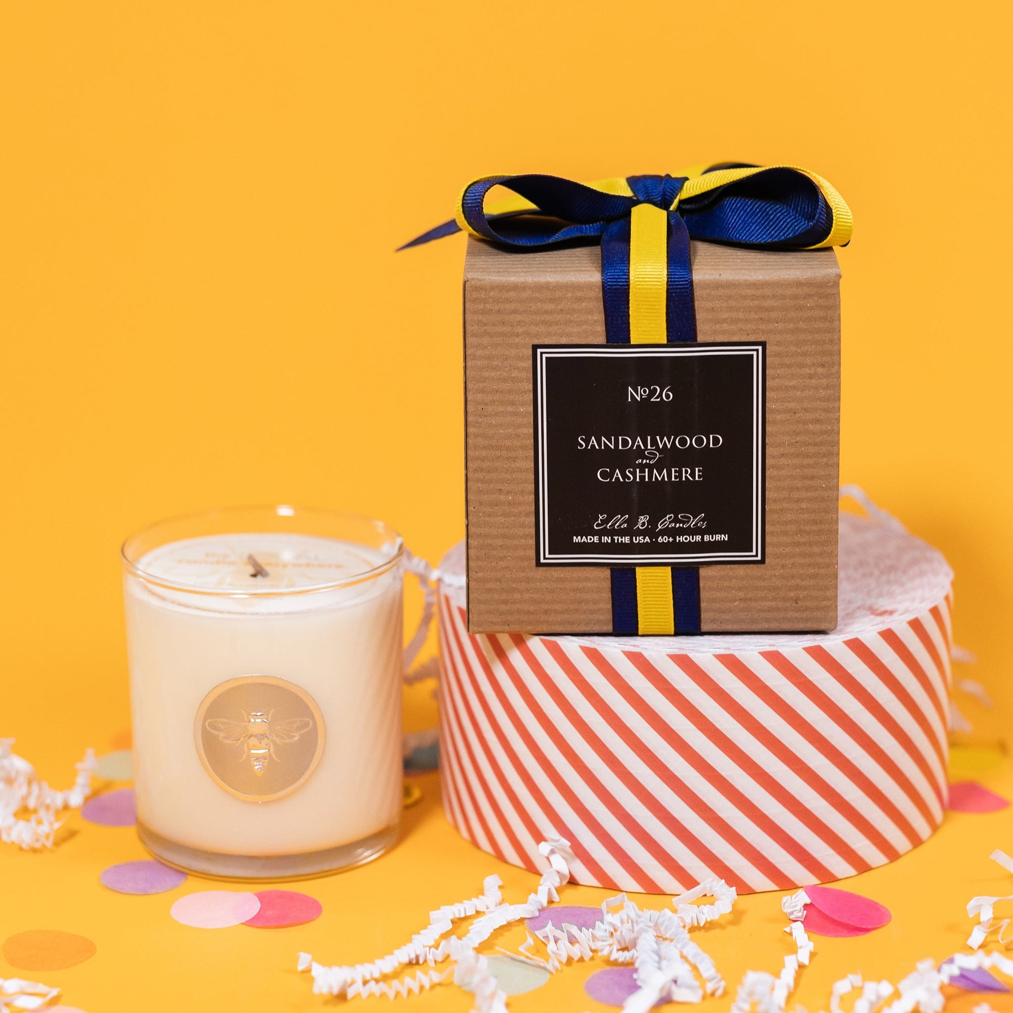 On a sunny mustard background is a candle with the back of a box and white crinkle with big, colorful confetti scattered around. The clear glass candle has a gold foil round label with an illustration of a bee on it. There is a kraft box with yellow and navy striped ribbon tied at the top and on the back is a square black label that says "No. 26 Sandalwood and Cashmere, Made in the USA, 60+ Hour Burn."