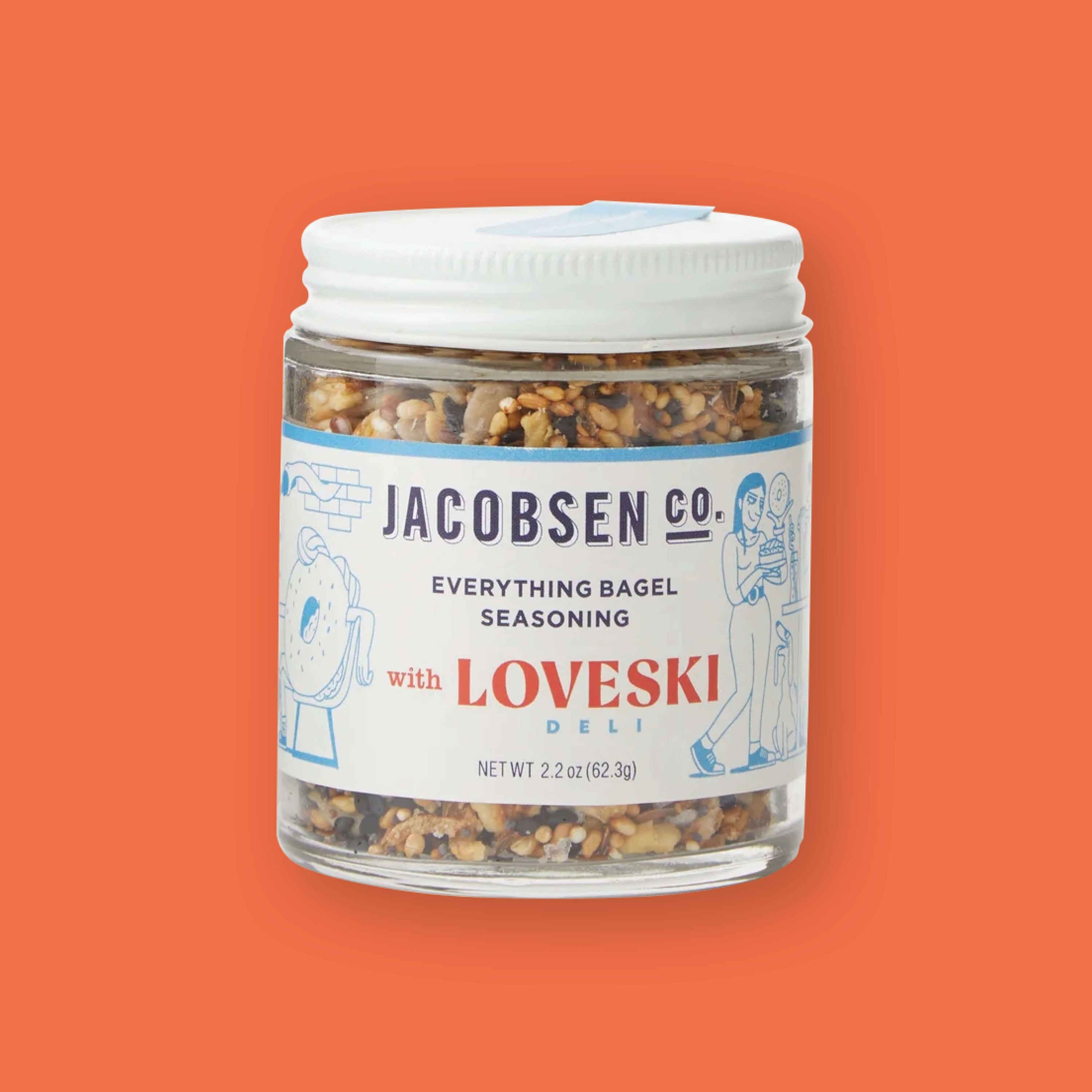 On an orangey-red background sits a jar. The clear jar has a white lid and label and shows various seasonings inside. It says "JACOBSEN CO. EVERYTHING BAGEL SEASONING" in navy, all caps block font. It says "with LOVESKI" in red font and "DELI" in a turquoise, all caps block font. There are illustrations in turquoise of a bagel with arms and a woman holding a bagel. New wt 2.2 oz (62.3g)