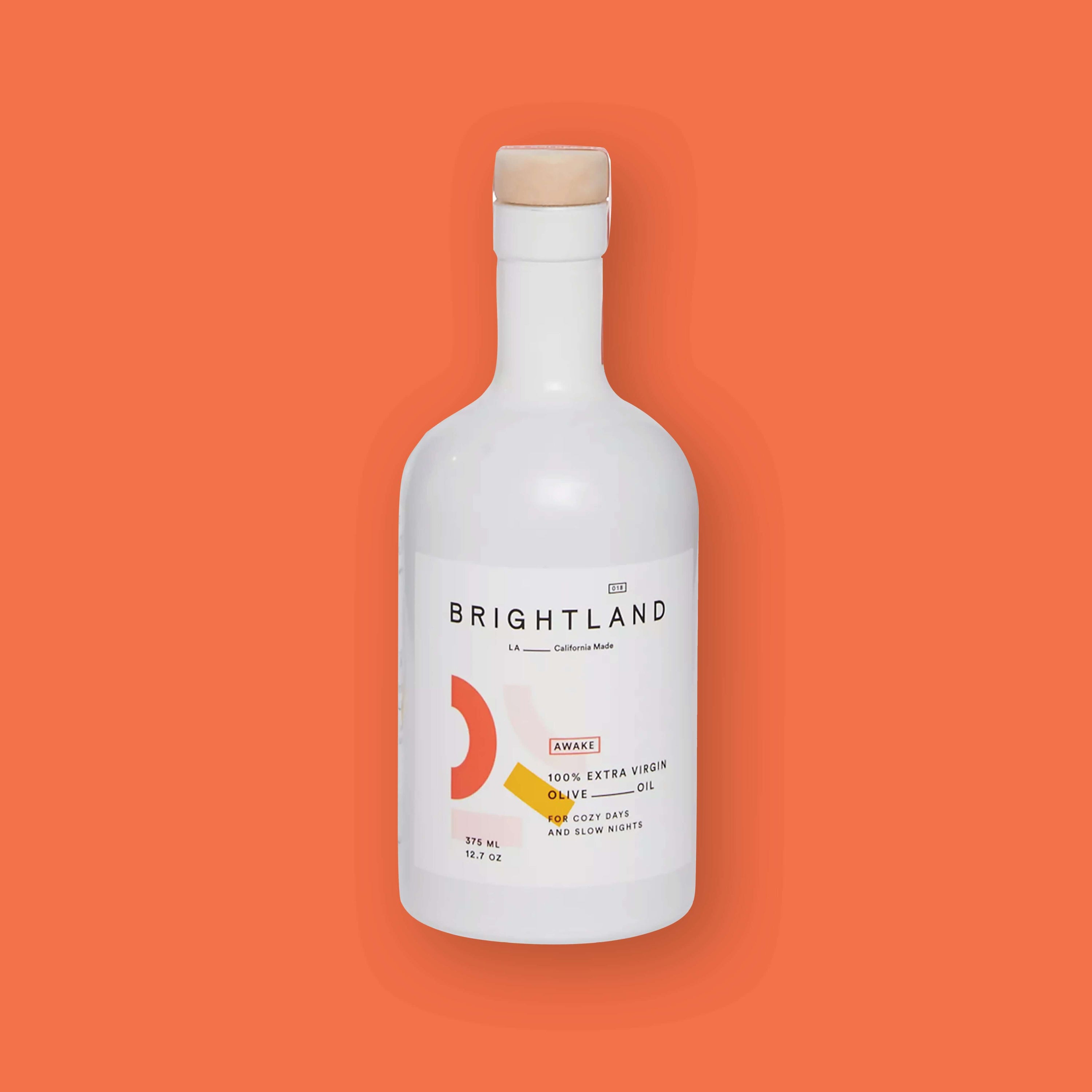 On an orangey-red background sits a bottle. This white bottle has a natural color top and white label. It says "BRIGHTLAND" in black, all caps block font and "LA_California Made" in black, block font. It says "AWAKE" in black, all caps block font inside an orangey-red rectangle and "100% EXTRA VIRGIN OLIVE_OIL FOR COZY DAYS AND SLOW NIGHTS" in black, all caps block font. It has two half circle outlines in orangey-red and pale pink. It also has two blocks in pale pink and mustard yellow. 12.7 oz 375 ml