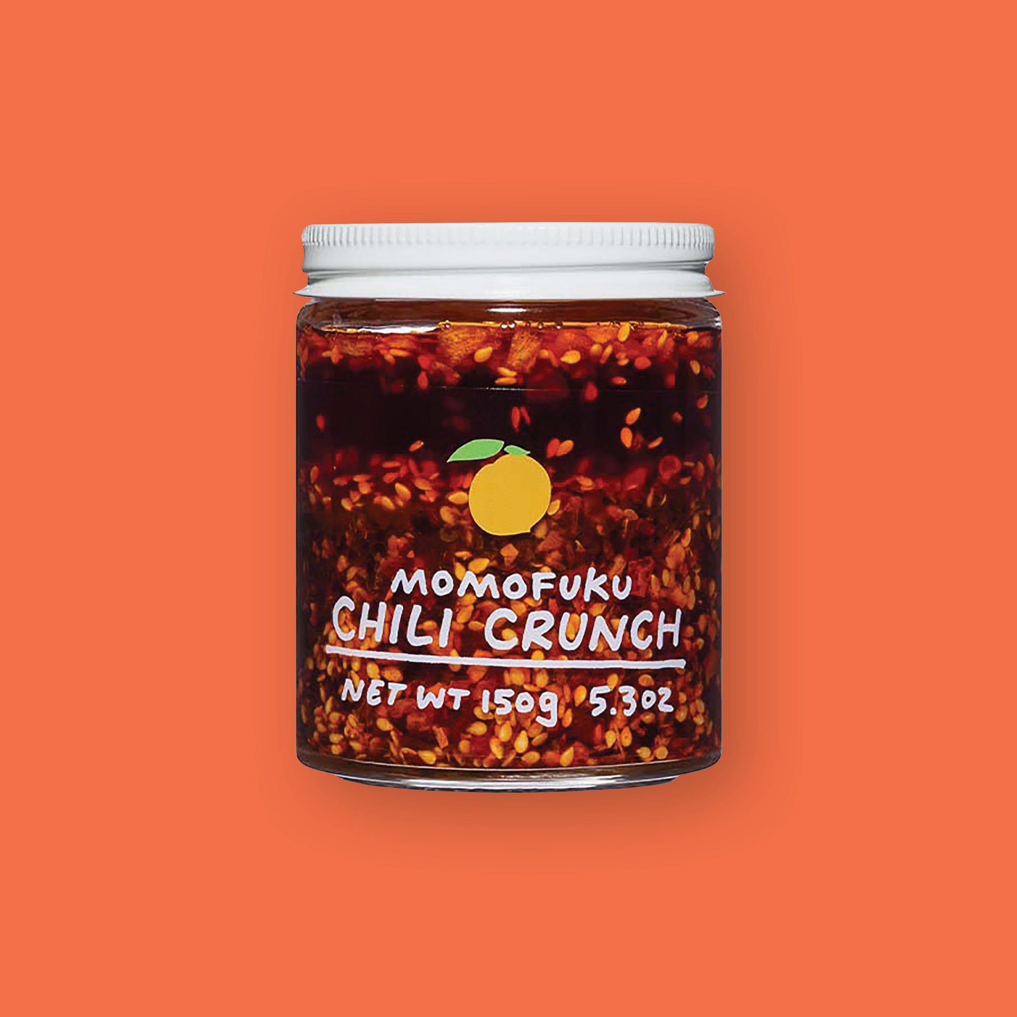 On an orangey-red background sits a jar. This clear jar has a white lid and is filled with chili seeds and liquid. On the front is an illustration of a yellow lemon and it says in white "MOMOFUKU CHILI CRUNCH" underlined in handwritten lettering. Net wt 150g 5.3oz