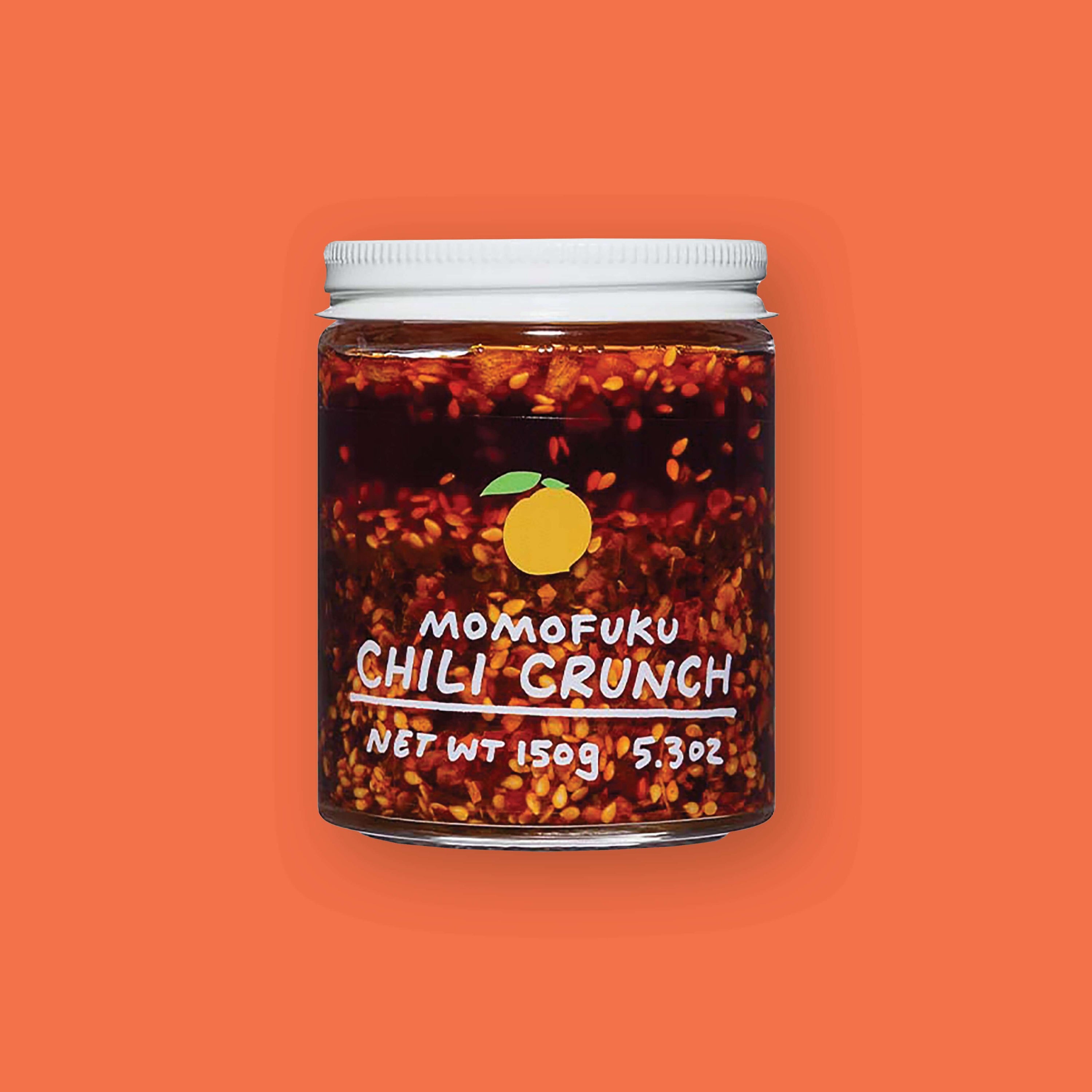 On an orangey-red background sits a jar. This clear jar has a white lid and is filled with chili seeds and liquid. On the front is an illustration of a yellow lemon and it says in white "MOMOFUKU CHILI CRUNCH" underlined in handwritten lettering. Net wt 150g 5.3oz