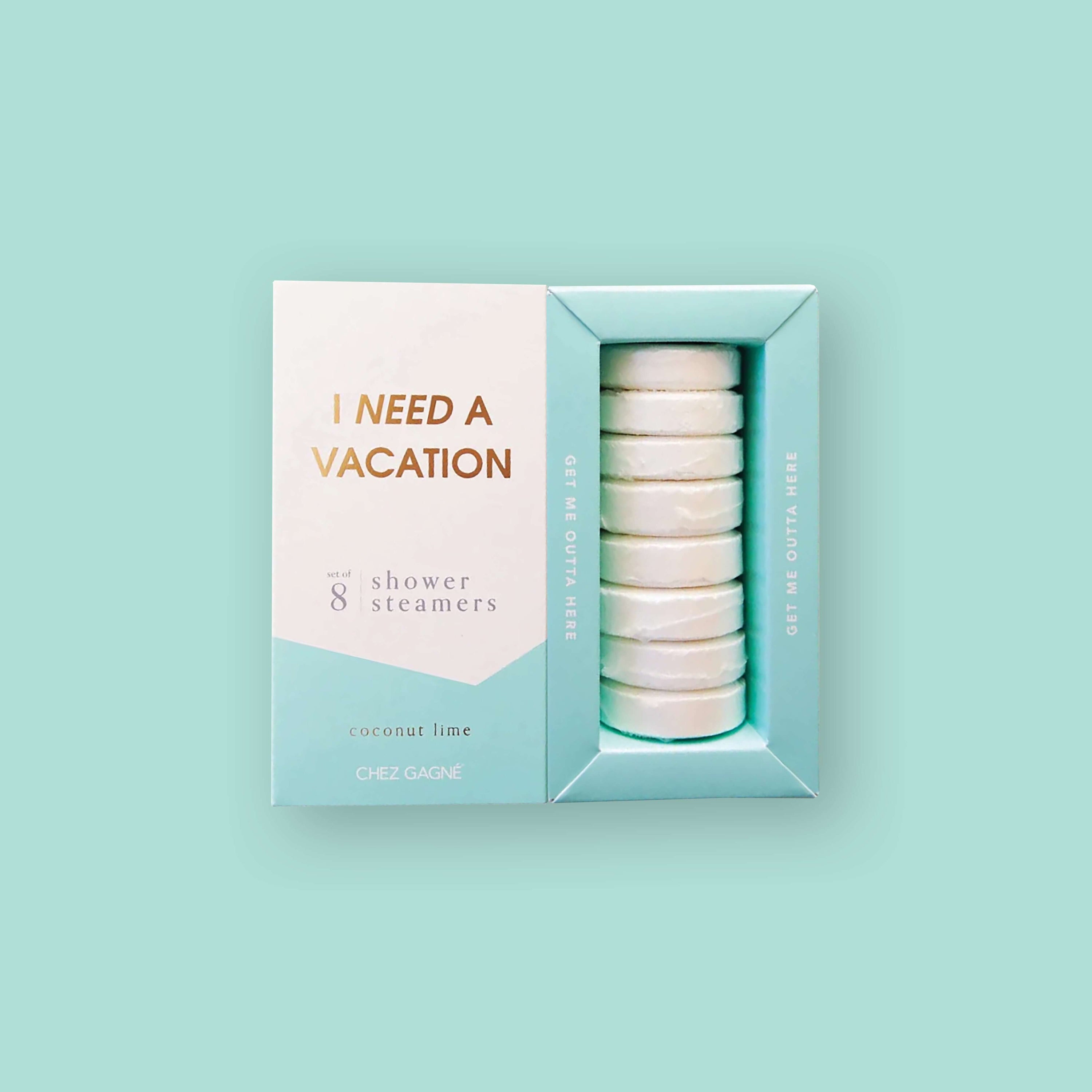 On a mint background sits two boxes. This picture is a close-up of a white and pool blue package that says "I NEED A VACATION" in gold foil, all caps block lettering. Under it says "set of 8" and " shower steamers" in grey, lowercase serif font. At the bottom it says "coconut lime" in gold foil, lower case serif font. To the right of it is a pool blue box with white shower steamers in it.