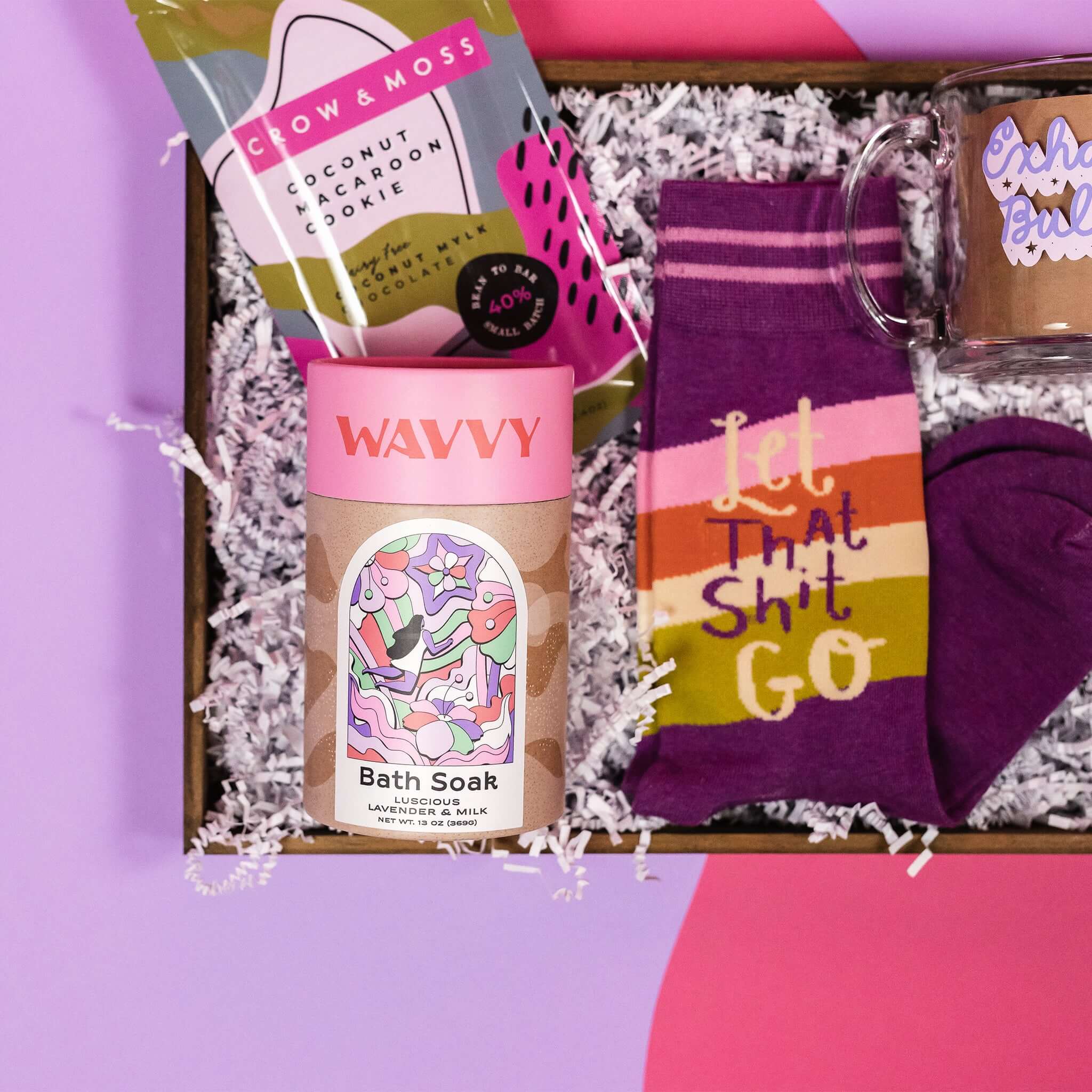On a hot pink and purple background sits a wooden tray with which paper crinkle and a handful of attractive gifts in a purple color theme. This photo is a close-up of the Wavvy Bath Soak in Luscious Lavender and Milk wiht a cute illustrated label and pink lid and purple novelty socks that say "Let that Shit Go."