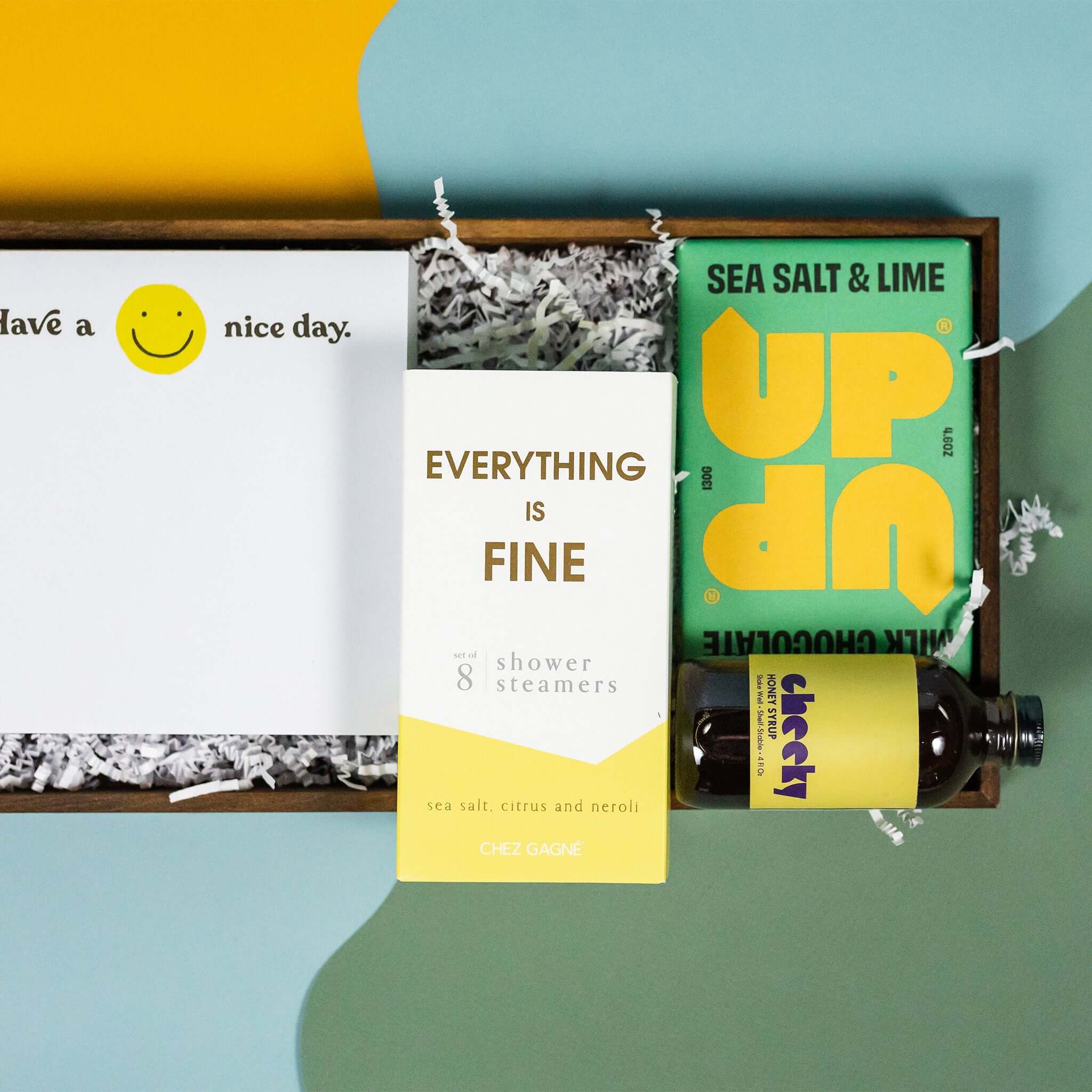 On a calming blue and green background with sunny yellow cutout sits a wooden tray of four products and white paper krinkle. Each product has some sunny yellow on the label. This is a close-up of a white square notepad with smiley face that says "Have a Nice Day," a pack of 8 Chez Gagne Sea Salt and Neroli "Everything is Fine" shower steamers, UP UP sea salt and lime milk chocolate bar, and mini bottle of Cheeky Honey Syrup.