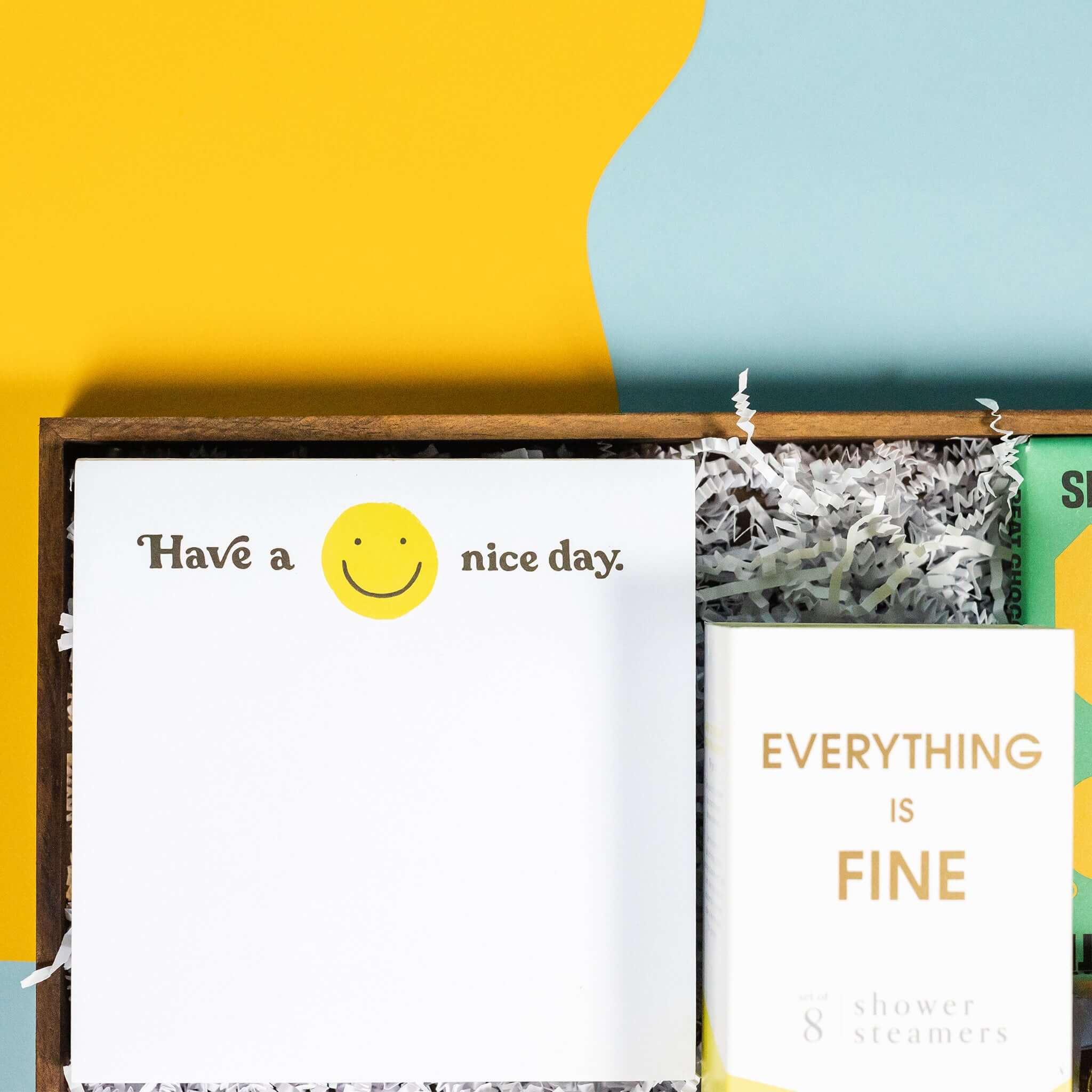 On a calming blue and green background with sunny yellow cutout sits a wooden tray of four products and white paper krinkle. Each product has some sunny yellow on the label. This is a close-up on the white square notepad with smiley face that says "Have a Nice Day."