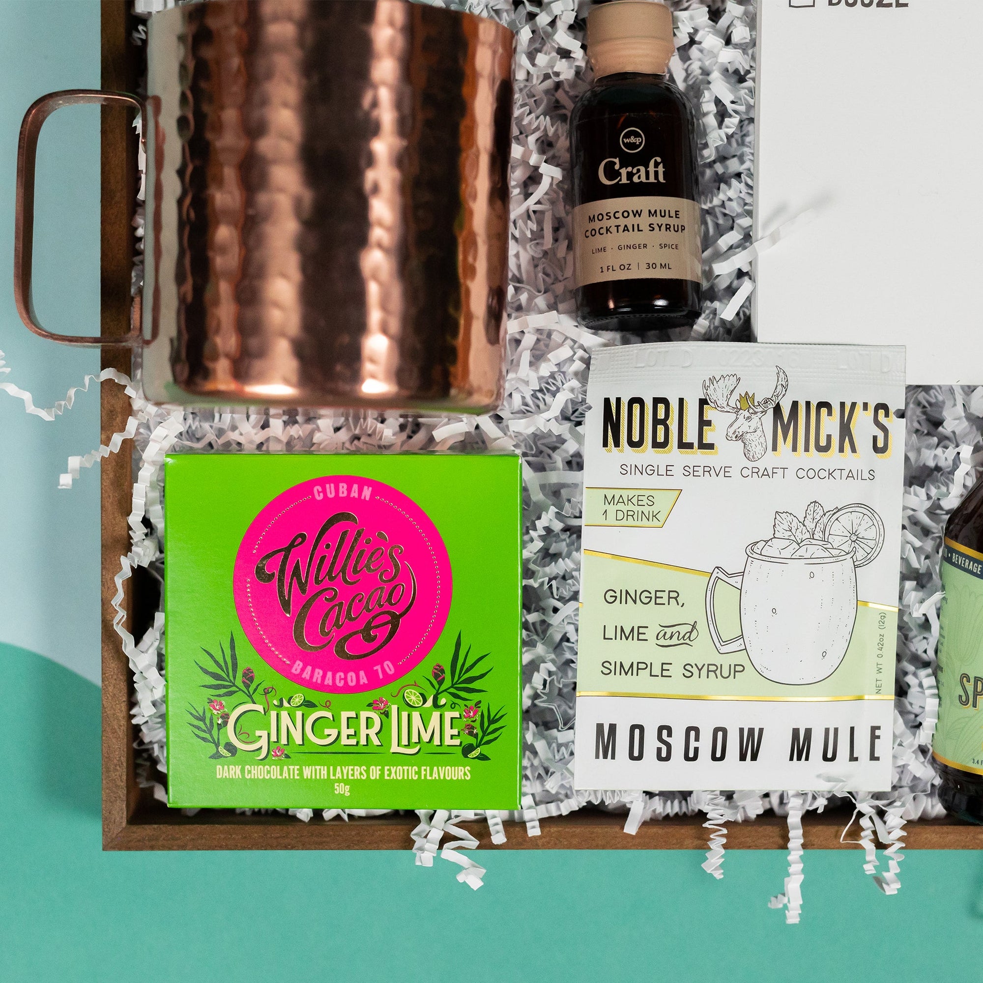 On a cool green and blue wavy background sits a wooden tray of moscow-mule themed gifts on white paper krinkle. This photo is a close-up on the hammered copper straight-sided mule mug, 1 oz mini bottle of W&P Moscow Mule Cocktail Syrup with lime, ginger and spice, Noble Mick's Single Serve Craft Moscow Mule Cocktail Mix and A 50g square of Willie's Cacao Ginger Lime Chocolate.