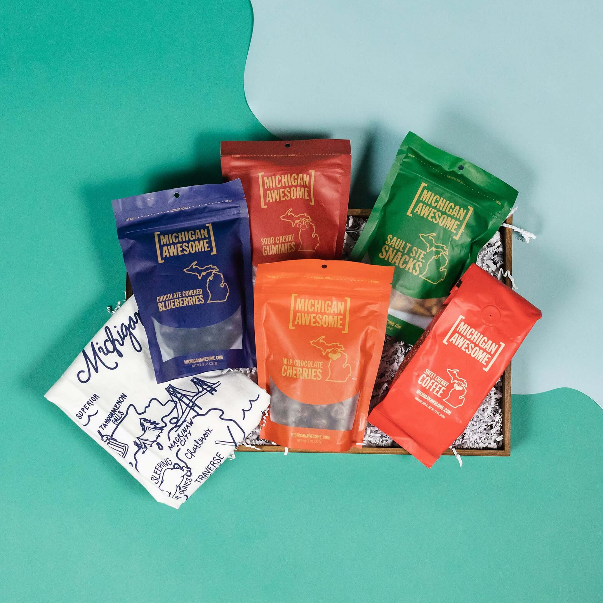 On a calming blue and teal background sits a wooden tray with an assortment of Michigan Awesome snacks and an illustrated map of Michigan tea towel.