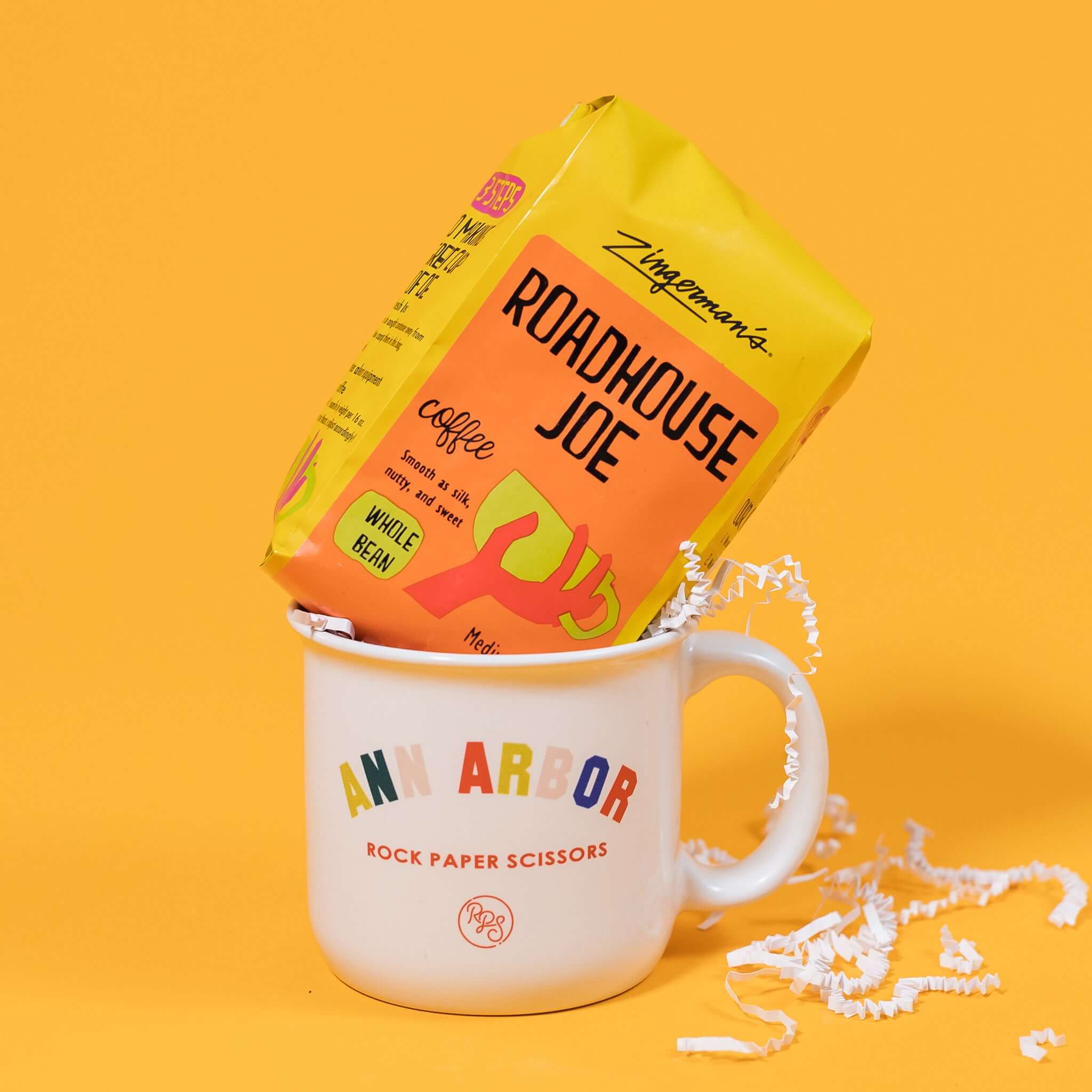 On a sunny mustard yellow and cool aqua backdrop sits a mug and a bag of coffee. The mug is a large white mug with colorful collegiate font that says "Ann Arbor" with "ROCK PAPER SCISSORS" below it. The bright yellow blue bag of coffee sits atop the mug. The coffee is Zingerman's brand Roadhouse Joe Blend. 'Smooth as silk and served daily at Zingerman's Roadhouse' Ground coffee. 12 oz (350 g). White crinkle overflows from the mug.