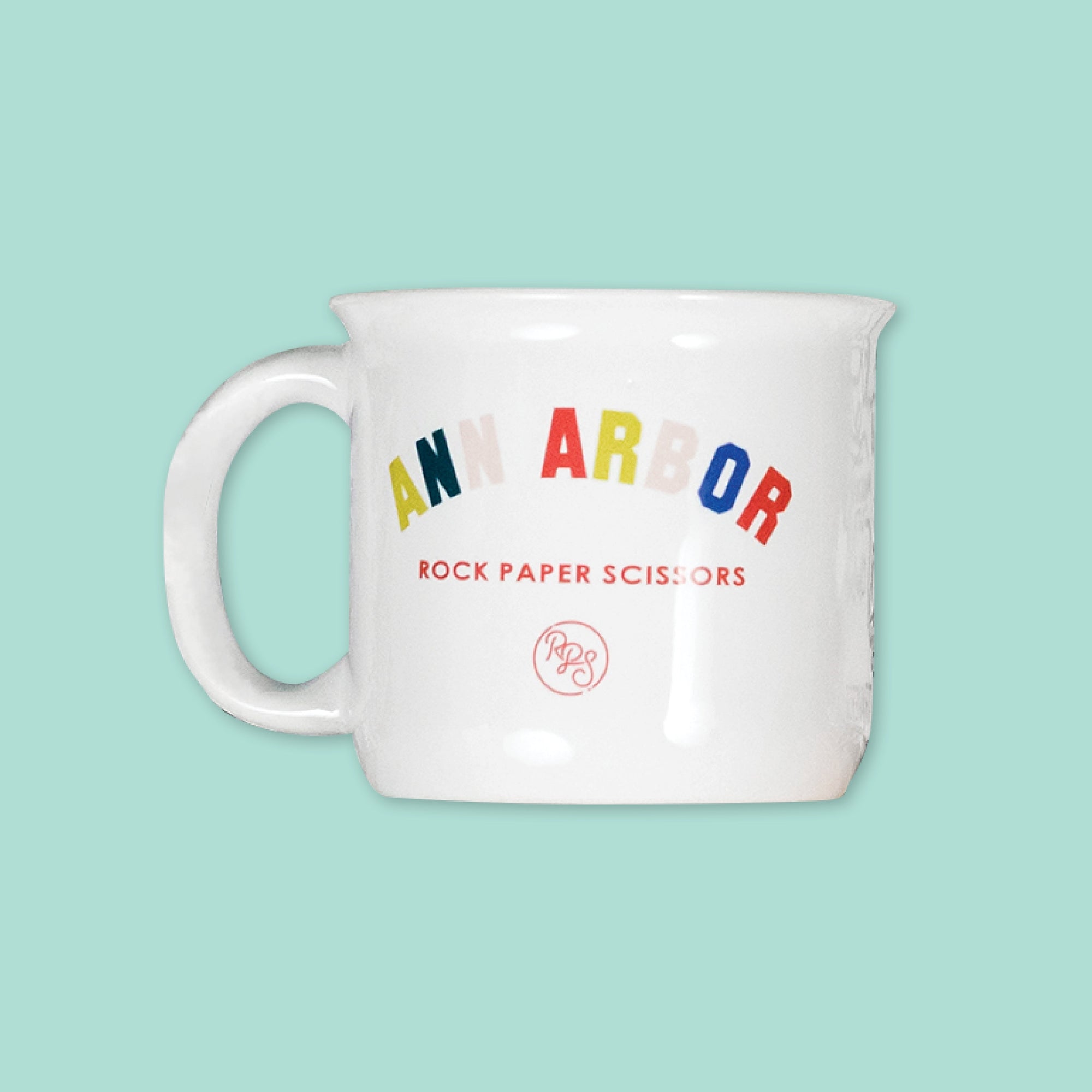 On a cool aqua background is a white mug with colorful collegiate font that says "Ann Arbor" with "ROCK PAPER SCISSORS" in red below it. 