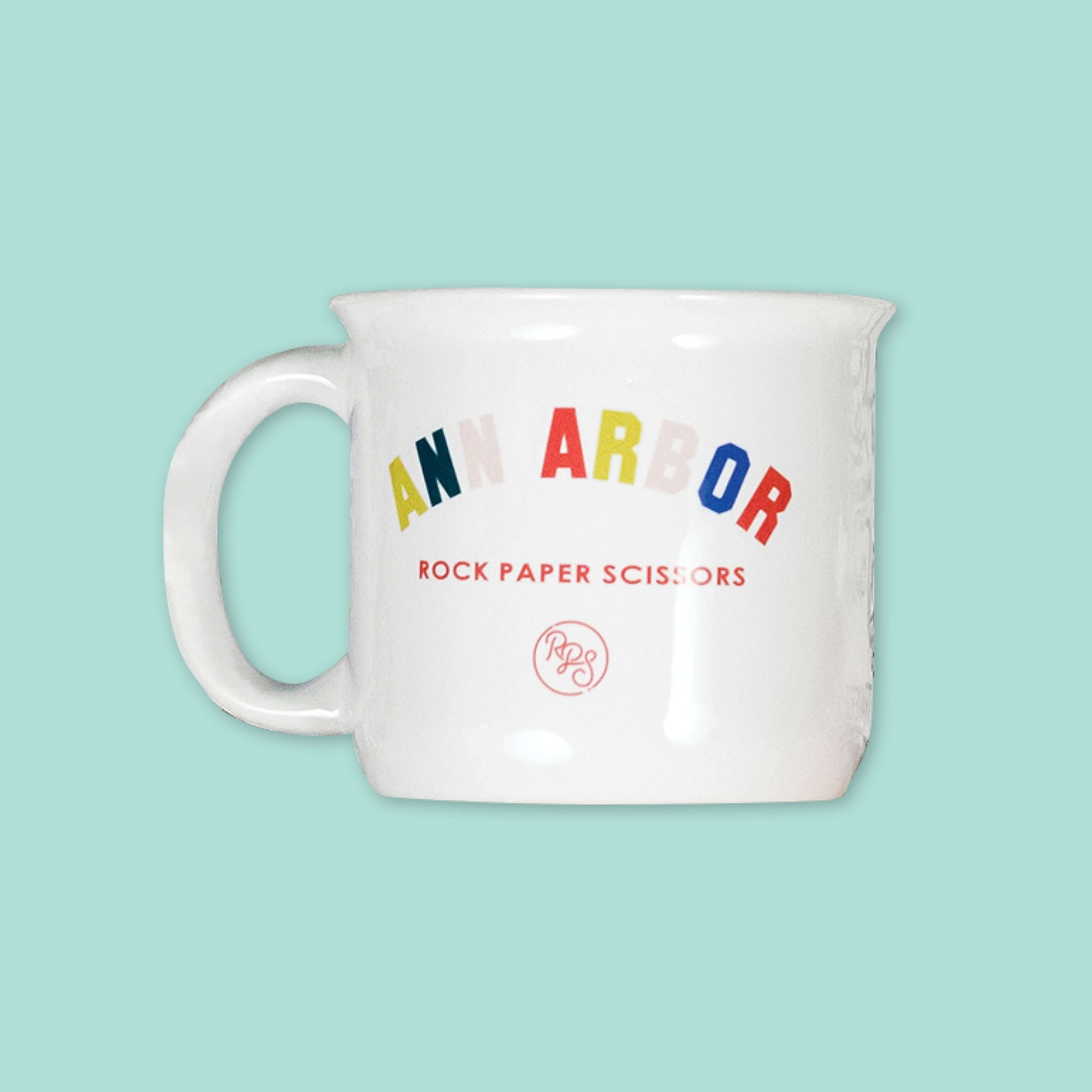 On a cool aqua background is a white mug with colorful collegiate font that says "Ann Arbor" with "ROCK PAPER SCISSORS" in red below it.