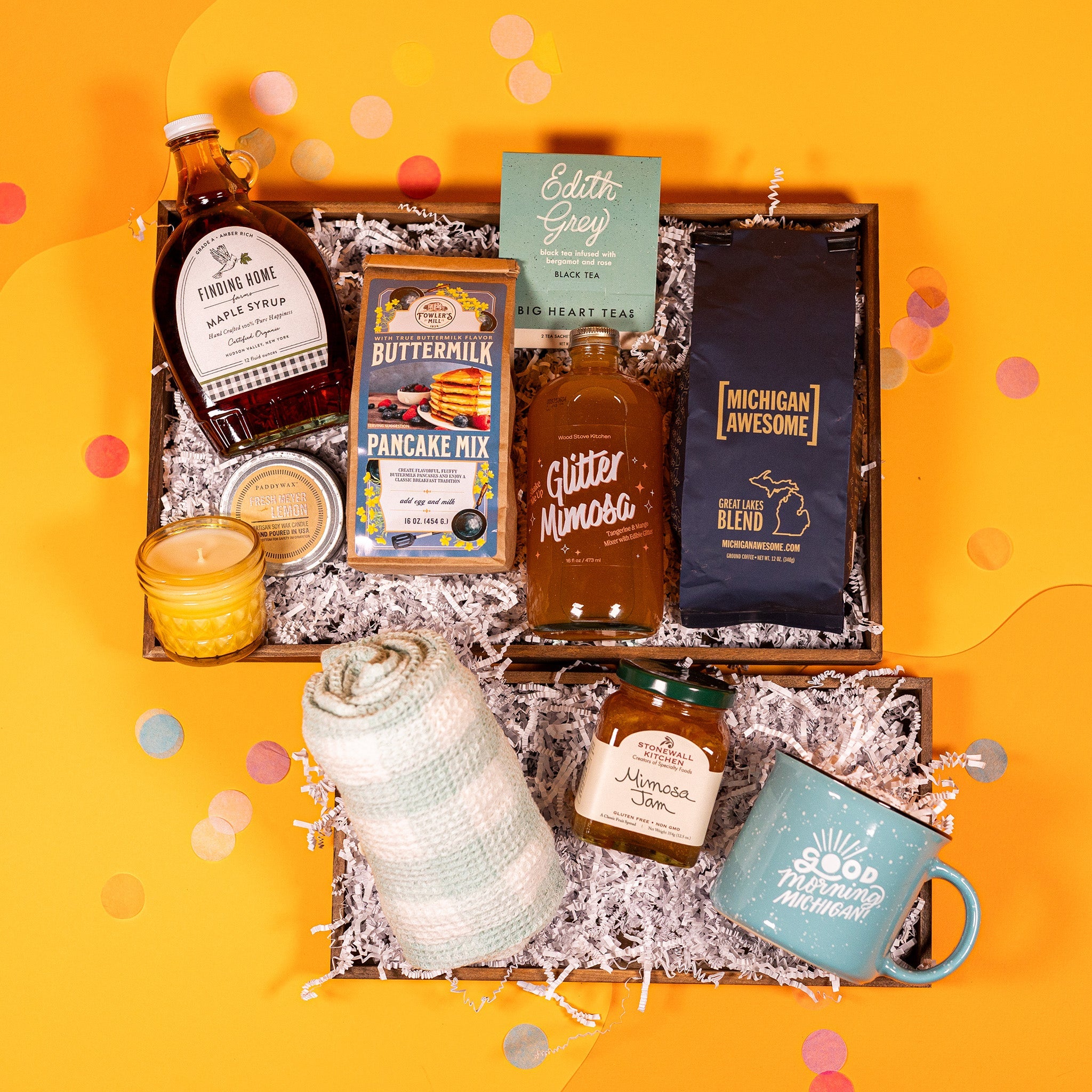 On a bright yellow background with coral pink confetti sits a collection of items on white crinkle paper for an Easter brunch gift box. A glass bottle of maple syrup, a yellow glass lemon candle, a brown paper bag of Fowler's Mill Pancake Mix, a teal and white box of Edith Grey Tea, glass bottle of Glitter Mimosa beverage, bag of Great Lakes Blend Coffee, a rolled up white and light teal striped tea towel, square glass jar of Mimosa Jam, and a light teal campfire mug that says "Good Morning Michigan."