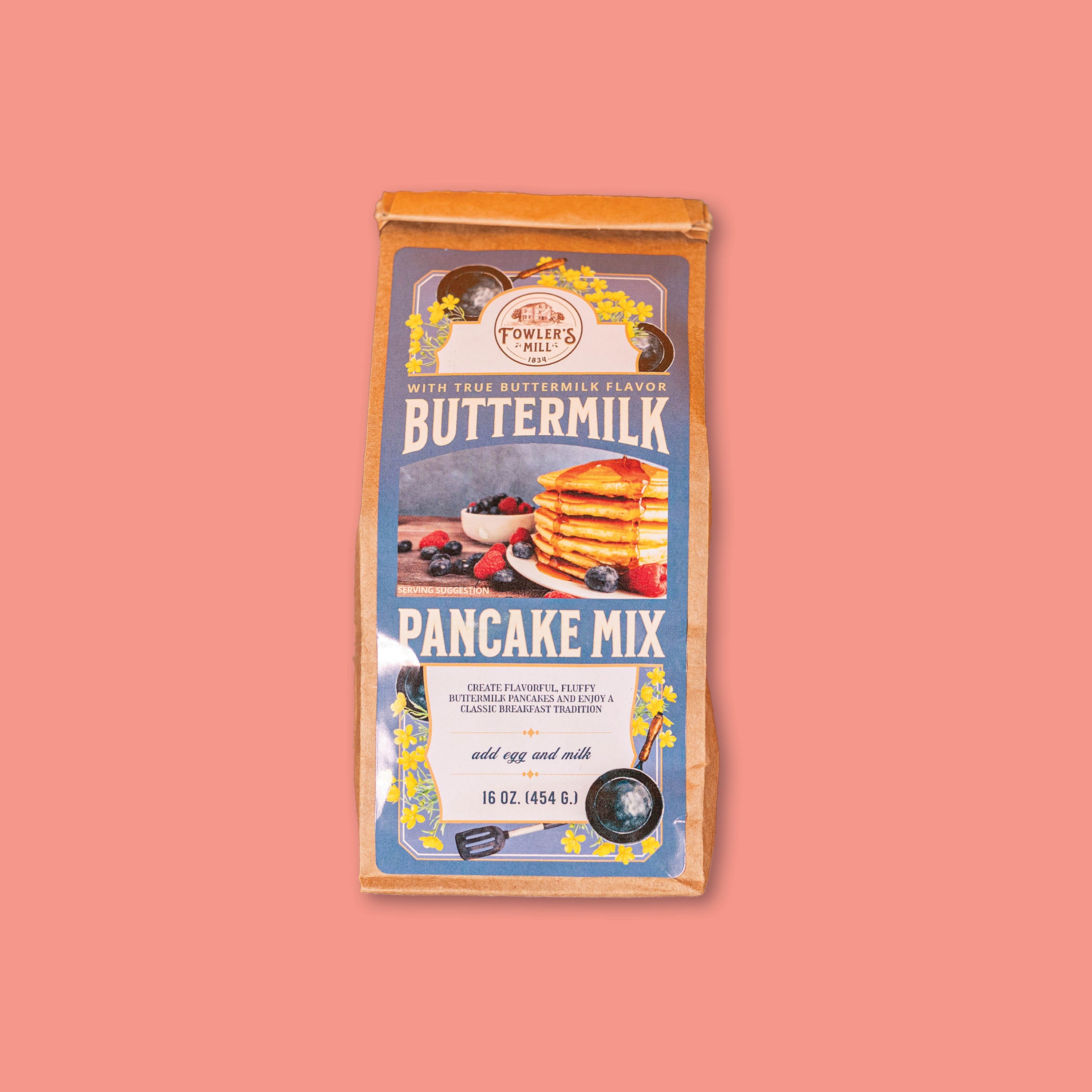 On a coral background sits a 16 oz. brown paper bag of Fowler's Mill Buttermilk Pancake Mix.