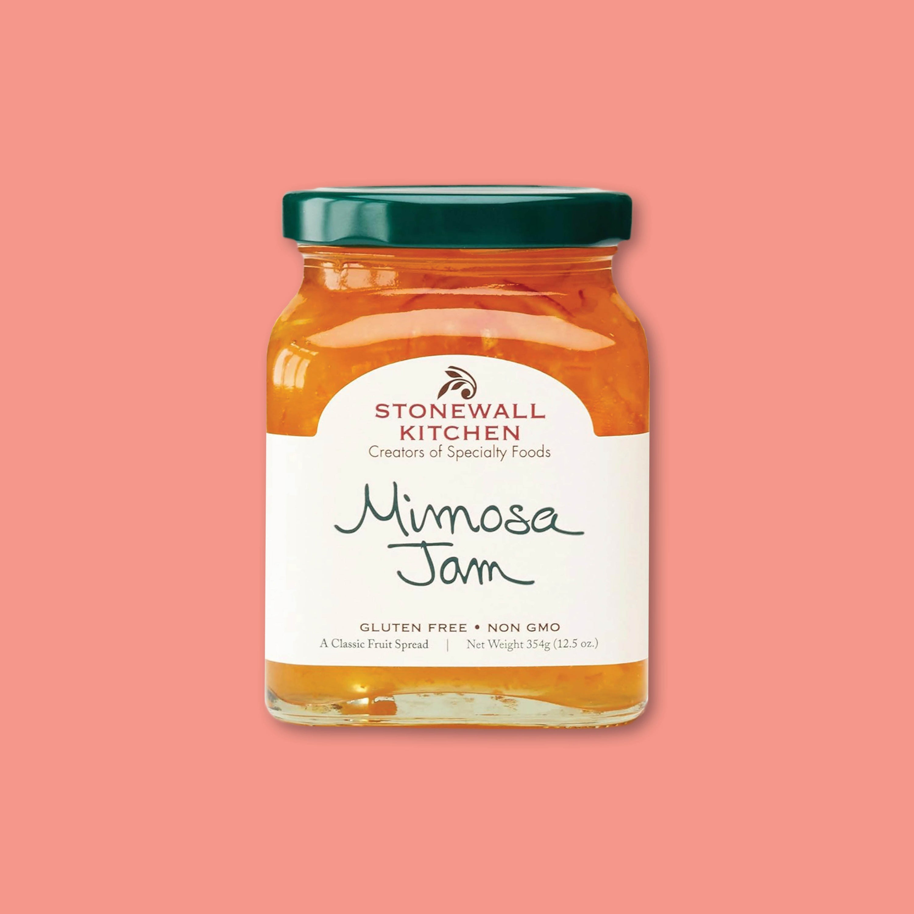 On a coral background sits a square 12.5 oz glass jar of Mimosa Jam from Stonewall Kitchen.