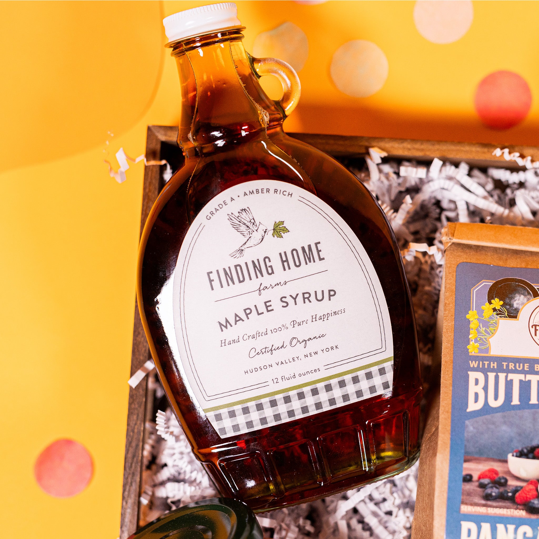 On a bright yellow background with coral pink confetti sits a gift box with white crinkle paper and a 12 oz. glass bottle of Finding Home Maple Syrup.