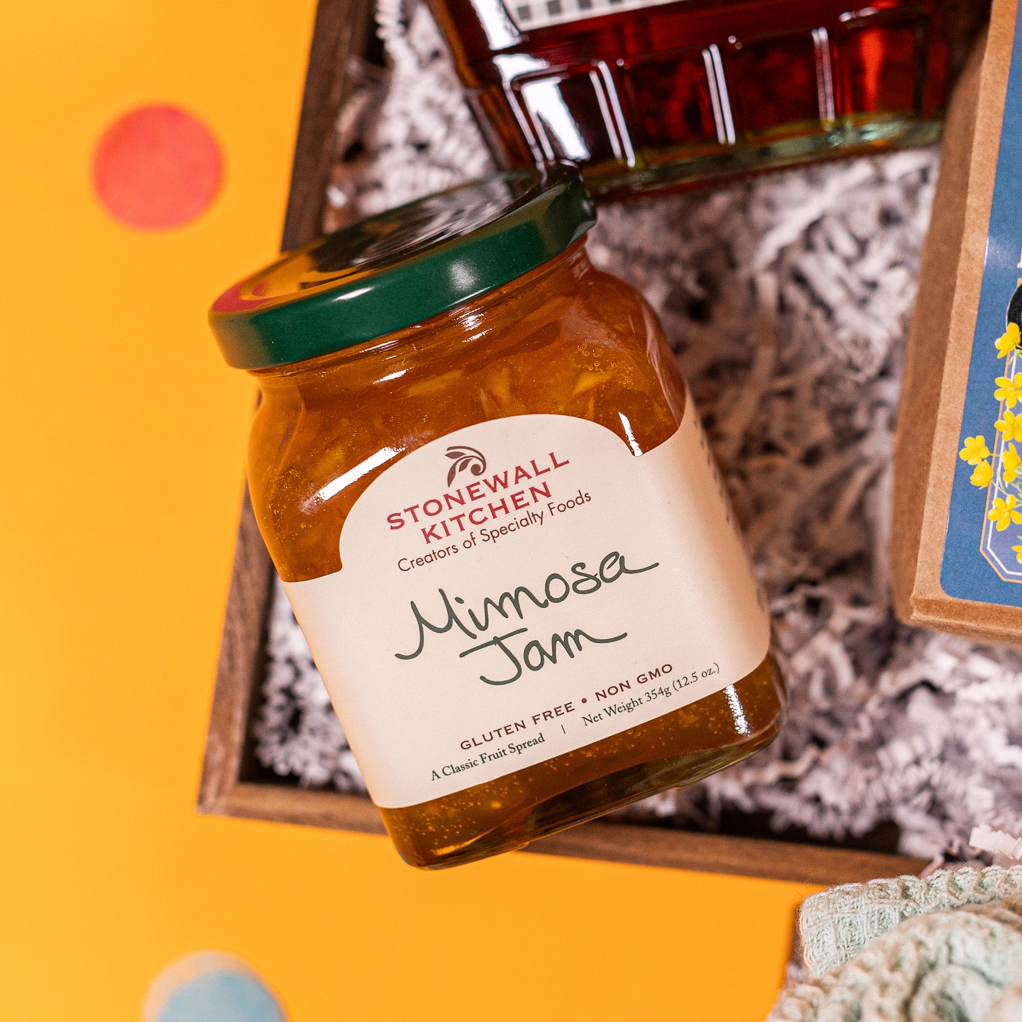 On a bright yellow background with coral pink confetti sits a gift box with white crinkle paper and a square 12.5 oz glass jar of Mimosa Jam from Stonewall Kitchen.